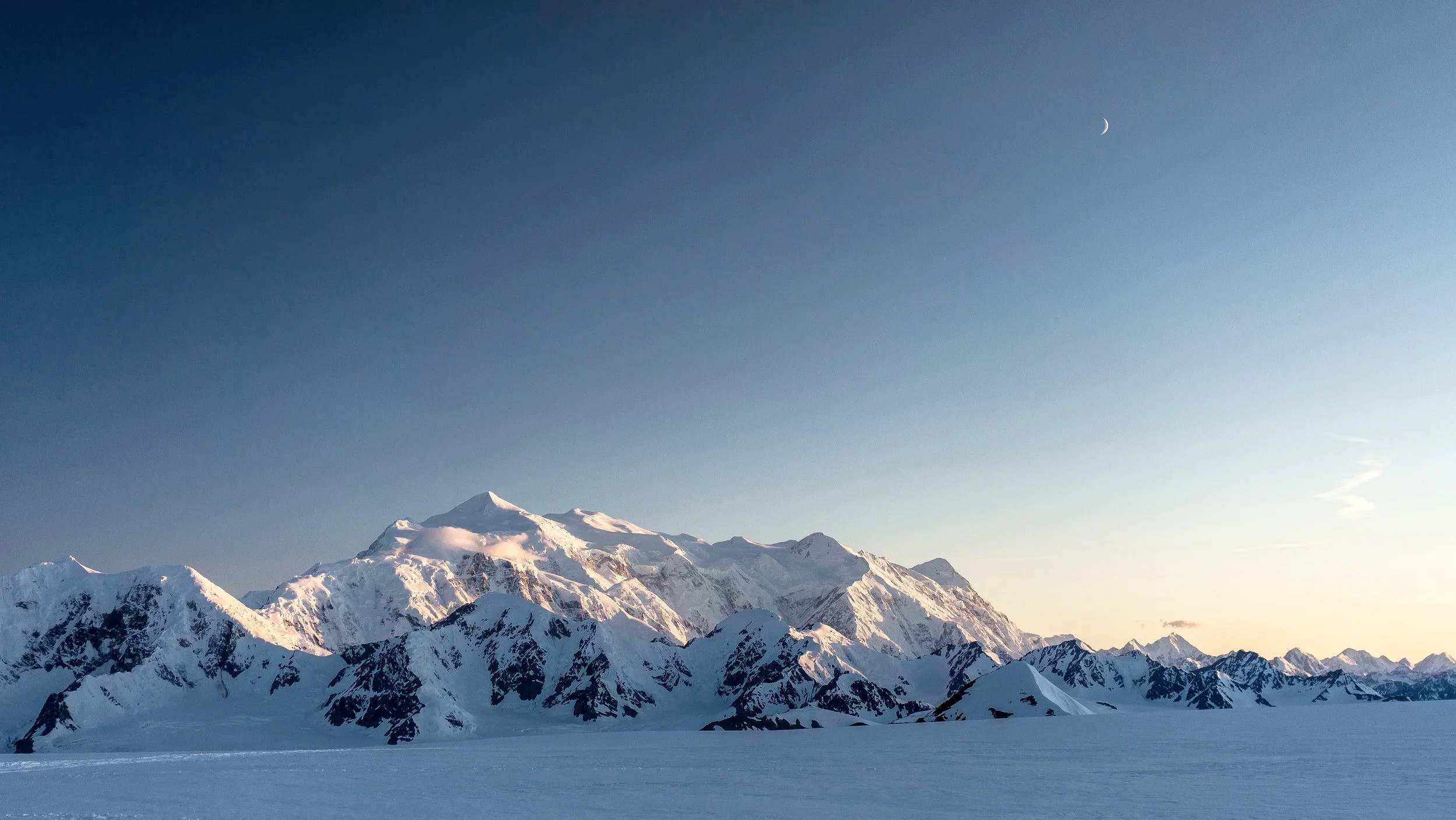 Snow-covered mountain range with a clear blue sky and a crescent moon above