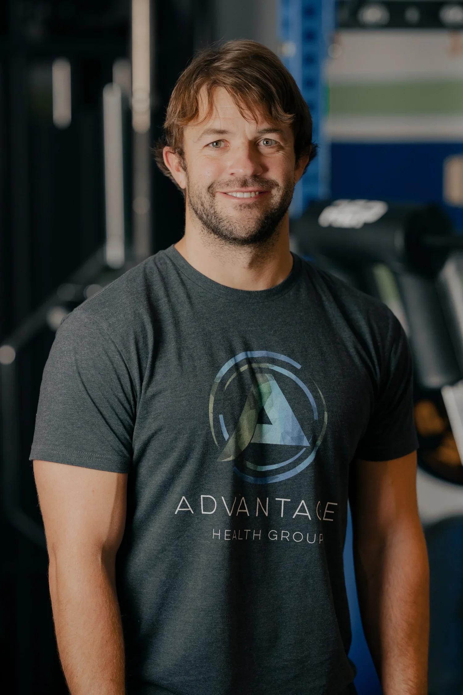 A smiling man standing in a gym, wearing a dark T-shirt with the logo 'Advantage Health Group'. Gym equipment can be seen in the background.