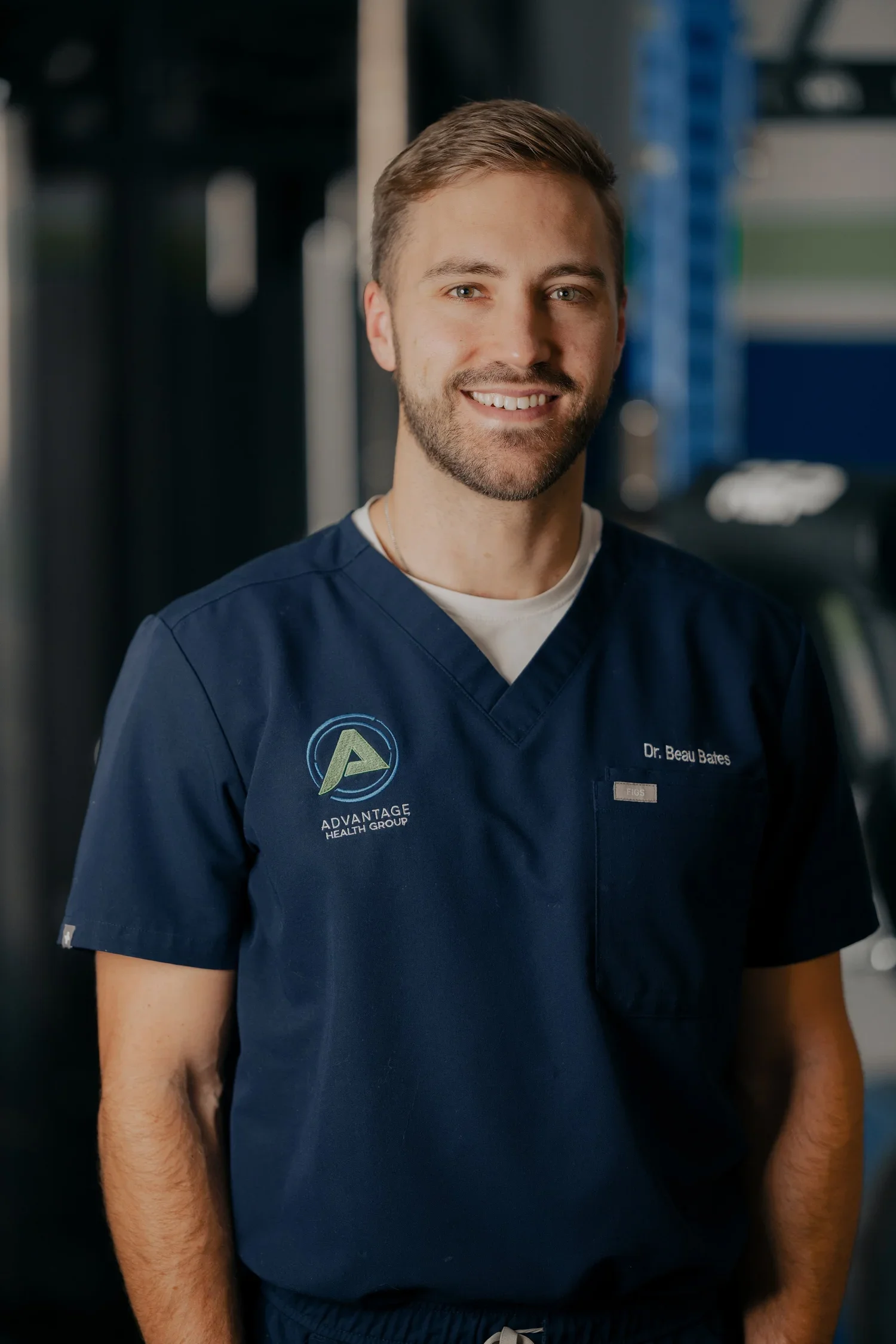 A smiling male healthcare professional wearing navy blue scrubs with a logo on the chest, standing in a medical or gym setting.