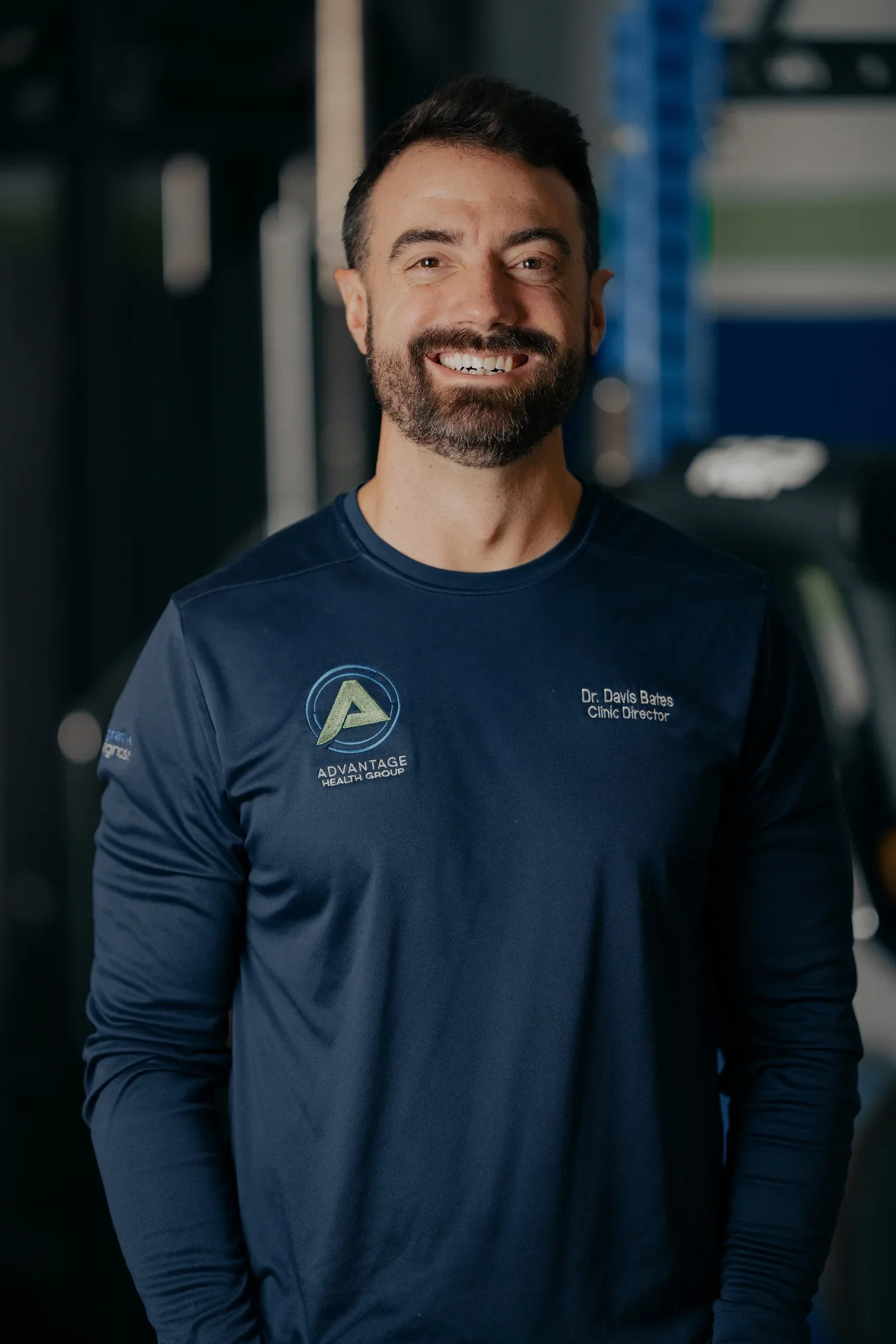 A smiling man with a beard and short dark hair, wearing a dark blue long-sleeve shirt with the logo and name of a health group and his title, standing in a gym with exercise equipment in the background.