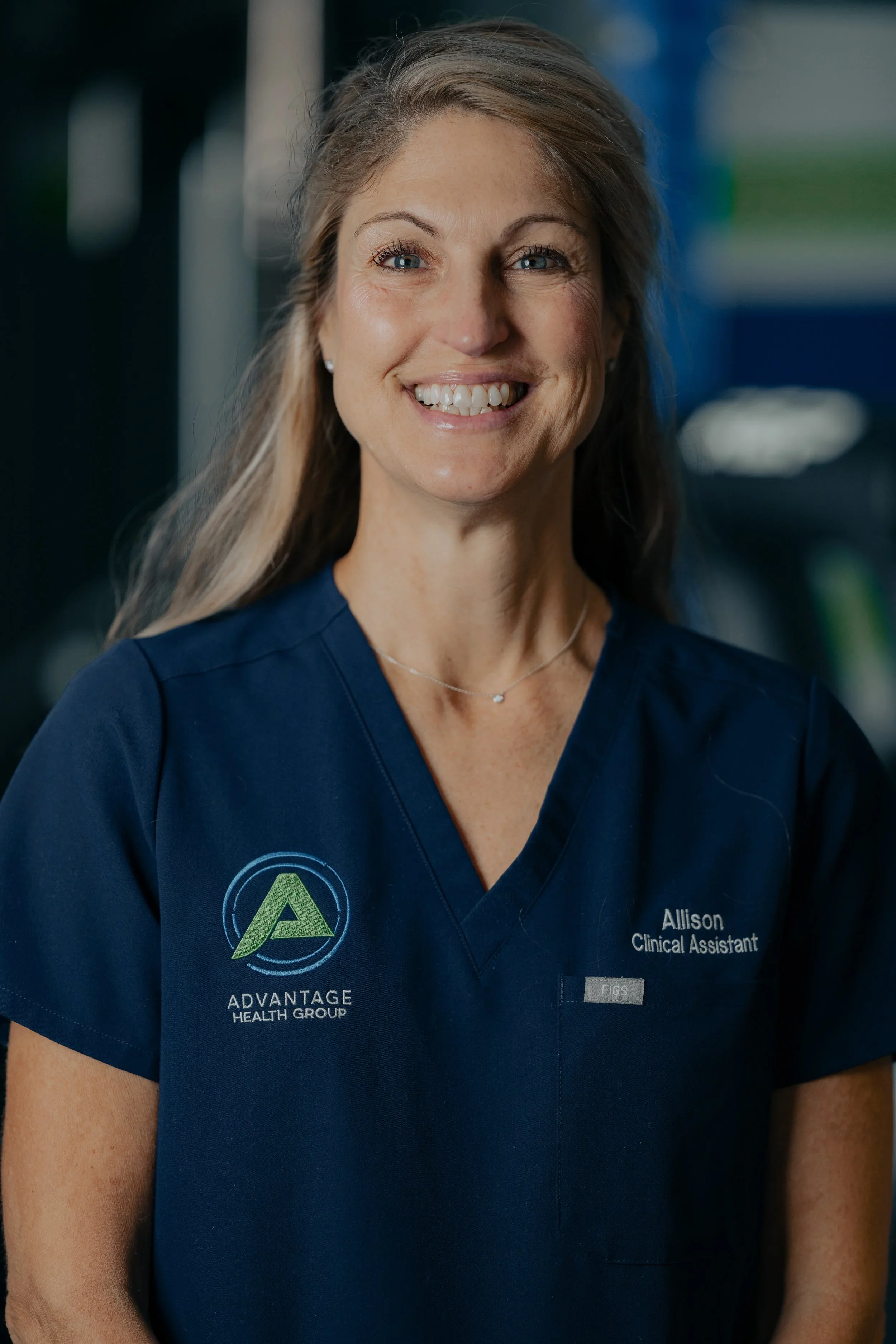 A woman with long light brown hair, blue eyes, and fair skin, smiling, wearing navy scrubs with 'Advantage Health Group' logo and her name tag, 'Allison, Clinical Assistant', on a blurred background.