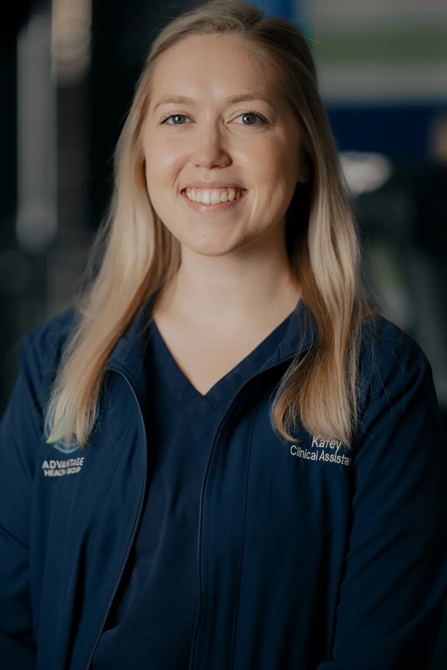 A smiling woman with long blonde hair, wearing a navy blue jacket with the words "Advantage Health Group" and "Karey, Clinical Assistant" visible, standing indoors.