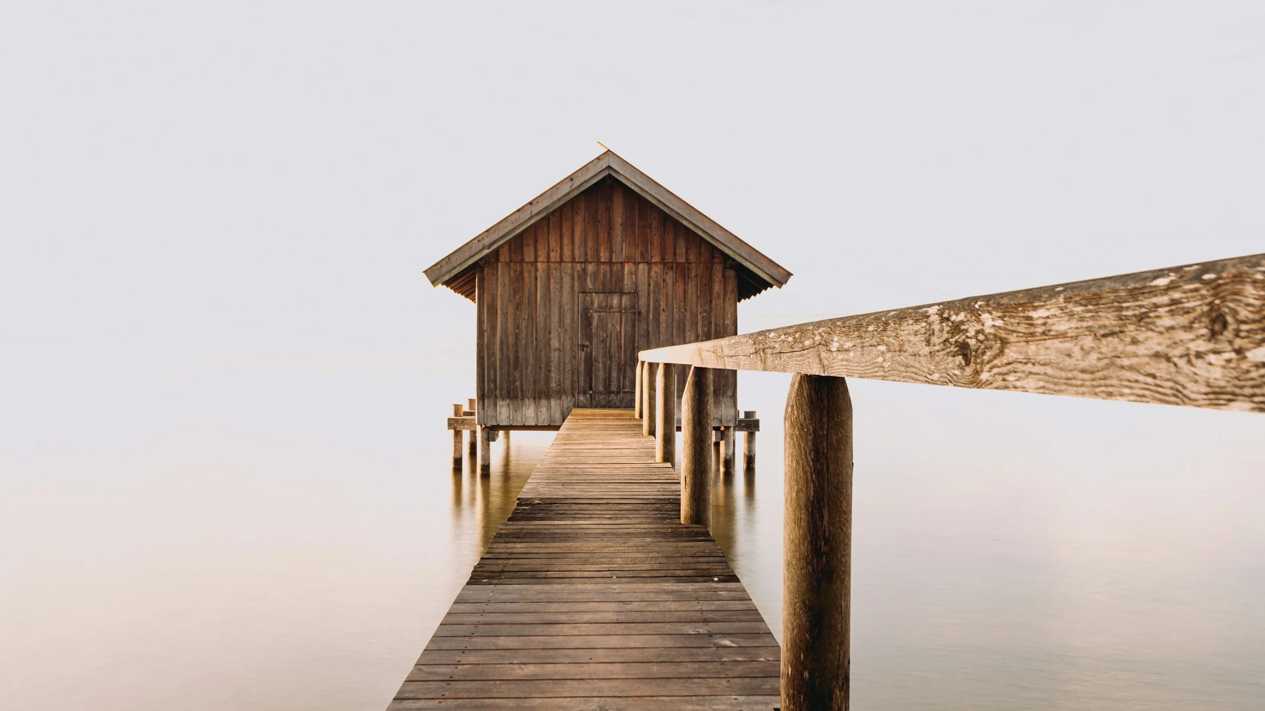 Wooden dock leading to a small wooden hut over water, with a wooden railing on the right side, surrounded by calm, overcast sky and water.