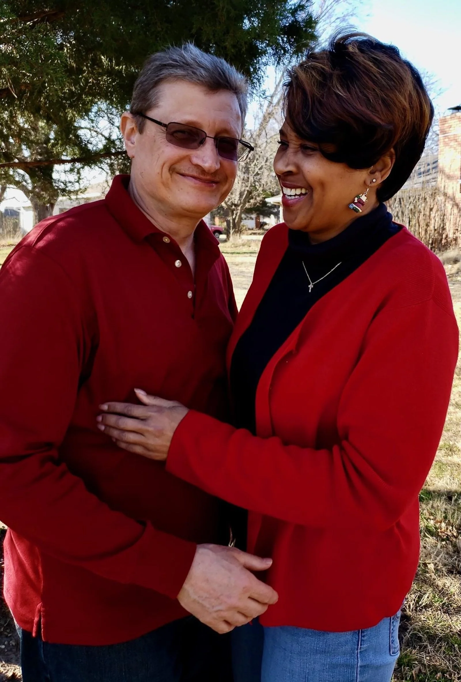 A smiling man with glasses and a woman with short dark hair and earrings, standing outdoors. The woman is wearing a red jacket, and the man is wearing a red shirt. They are embracing and appear happy. There are trees and buildings in the background.