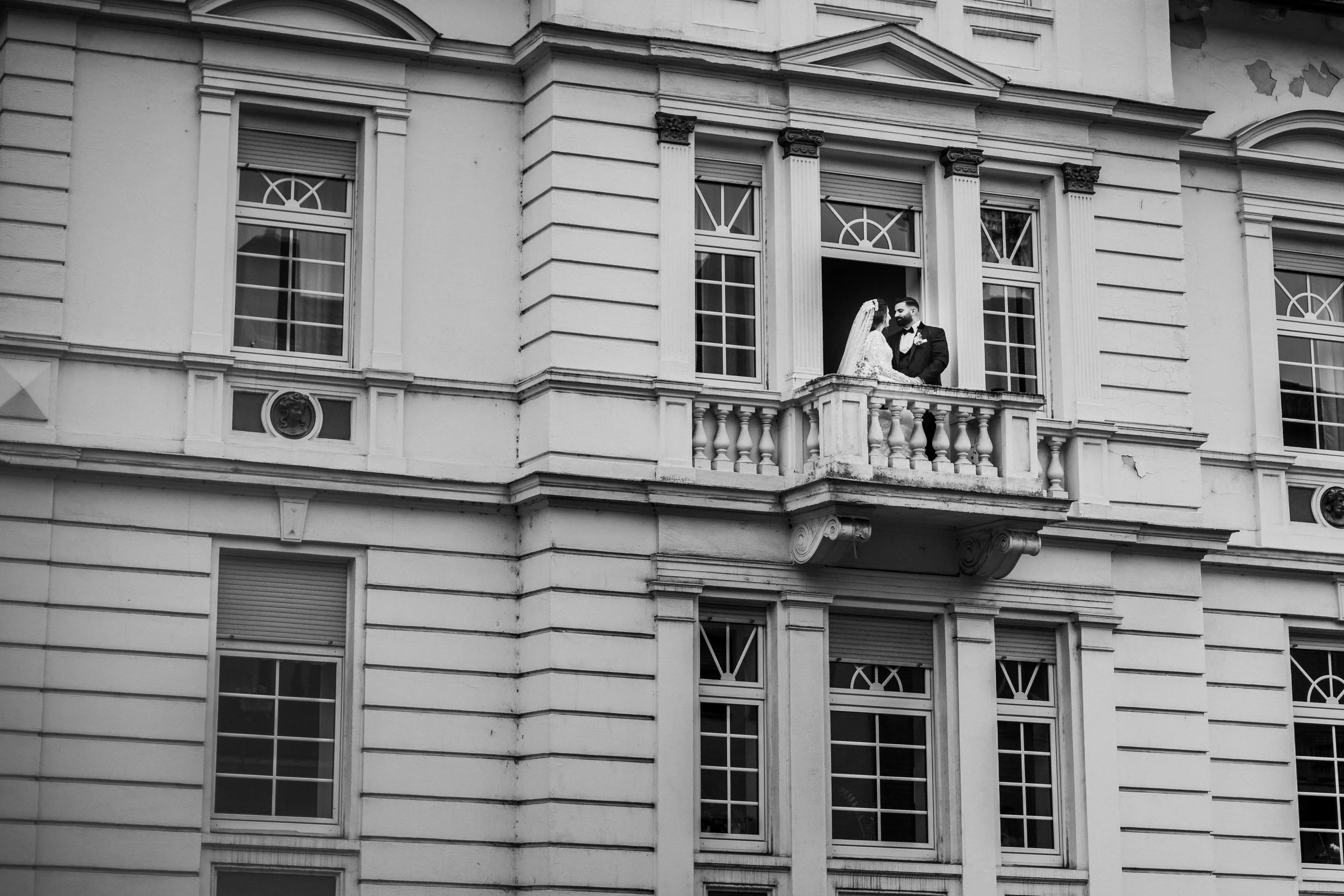Ein schwarzer und weißer Foto eines Hochzeitspaares auf einem Balkon eines alten Gebäudes, das Gebäude hat eine klassische Architektur mit mehreren Fenstern und Säulen.