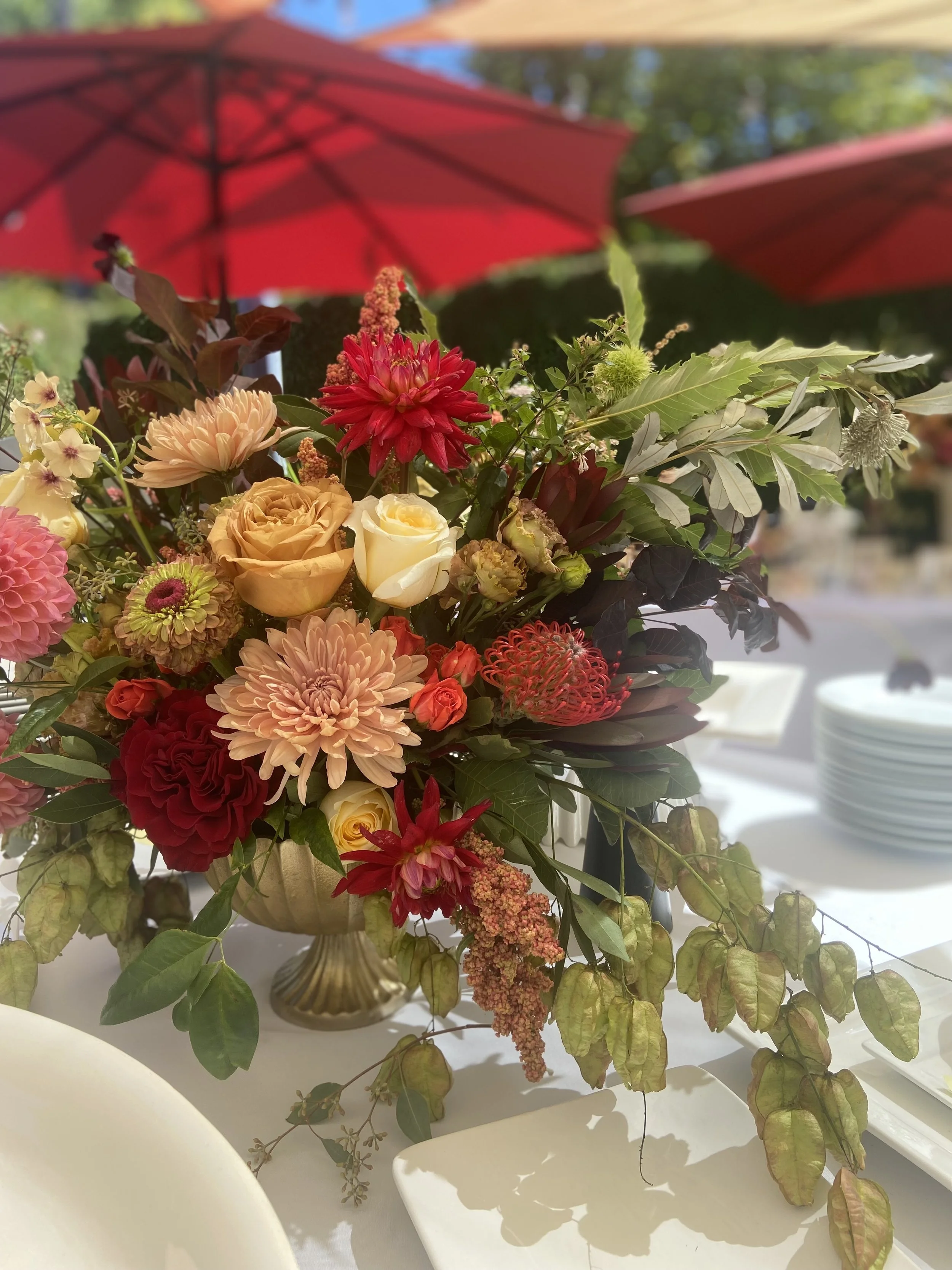 A floral centerpiece with various colorful flowers, green leaves, and a decorative gold vase, set on a table with white plates and a white tablecloth under red umbrellas outdoor.