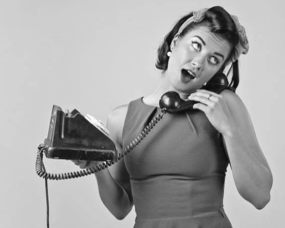 Woman with vintage hairstyle and dress talking on a corded landline phone, making a surprised or playful facial expression.