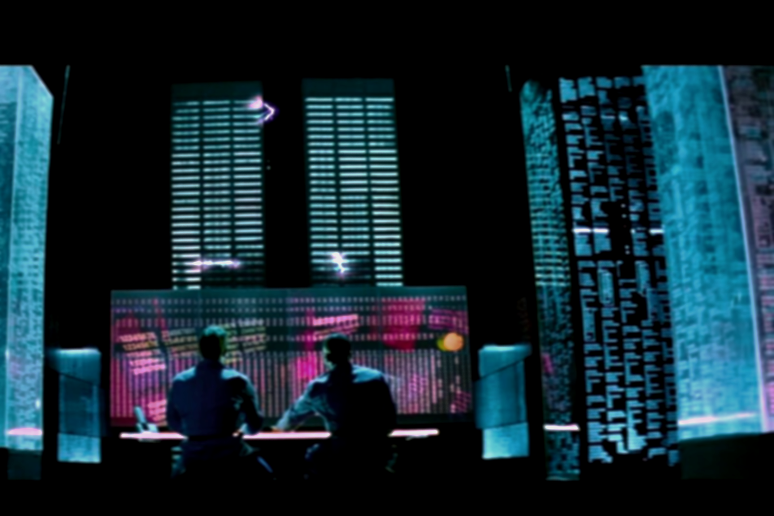 Two individuals sitting at a high-tech conference table, facing a large digital screen displaying vibrant graphics, in a dimly lit futuristic office.
