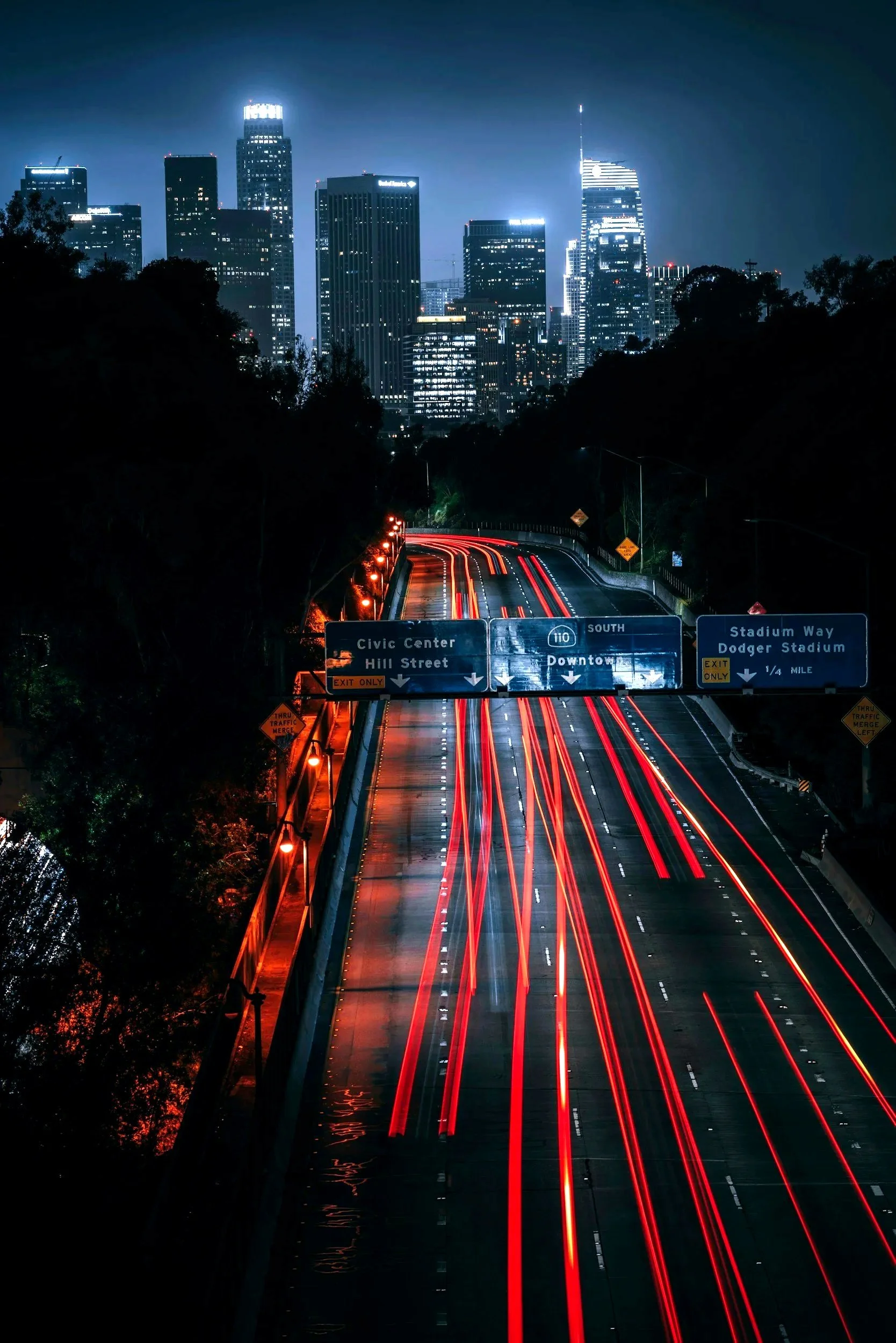 Nighttime cityscape with illuminated skyscrapers and streaks of red and white lights from moving vehicles on a highway, with traffic signs and trees along the road.