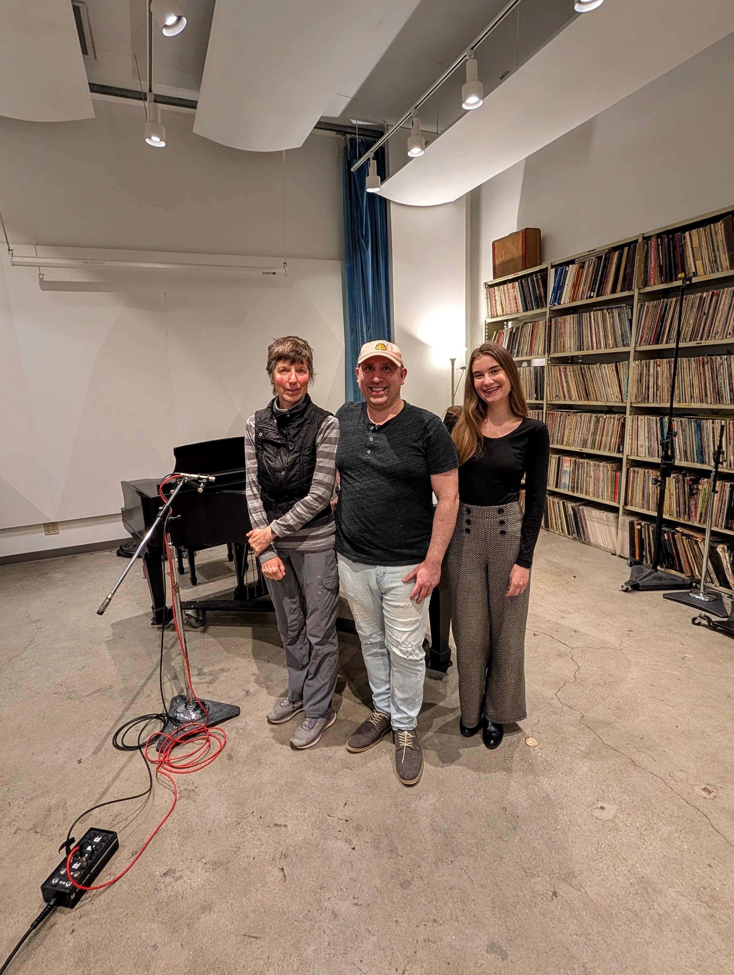 Three people standing next to a grand piano in a room with a bookshelf filled with records. They are smiling at the camera.
