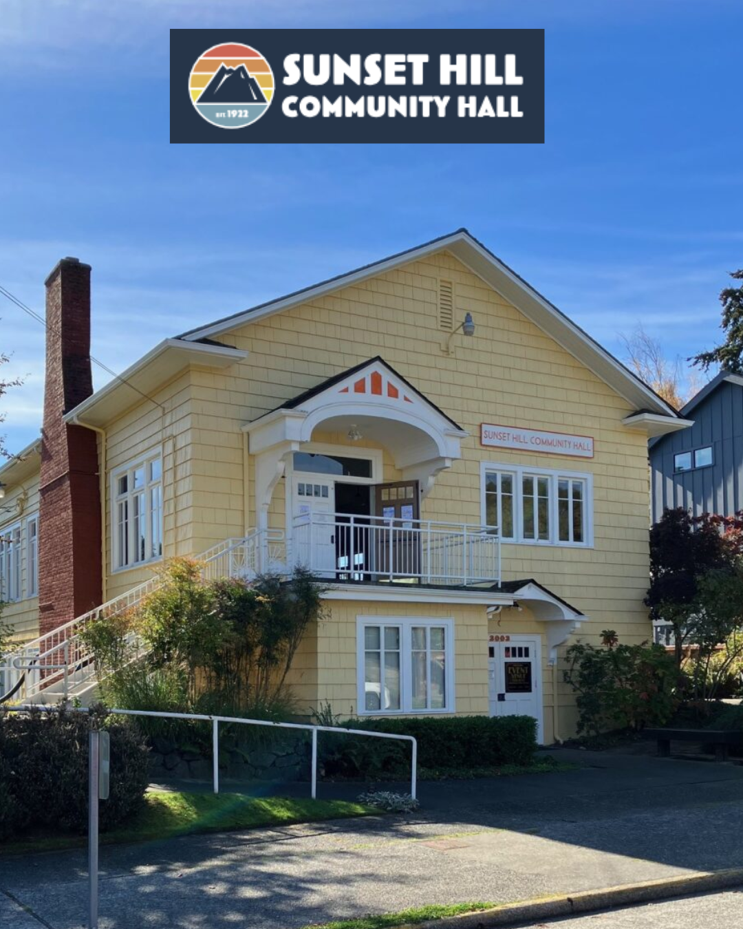 Yellow two-story building with white trim labeled as Sunset Hill Community Hall, featuring a small balcony with a white railing, white door, and multiple windows, with a sign above the balcony and a sign on the door.