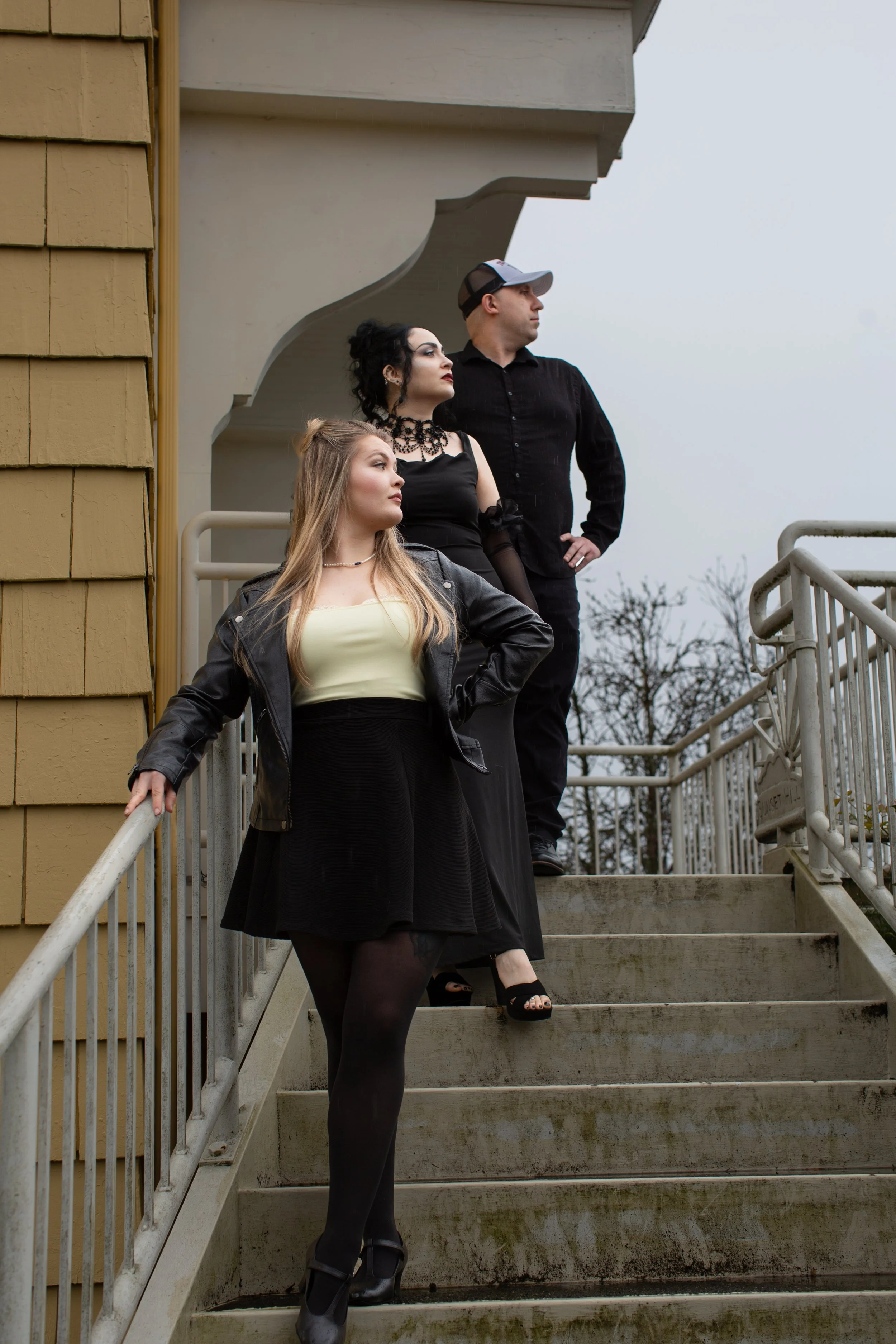 Three people standing on outdoor staircase, two women and one man, dressed in dark clothing with gothic style accessories, urban setting, cloudy sky, leafless trees in background.