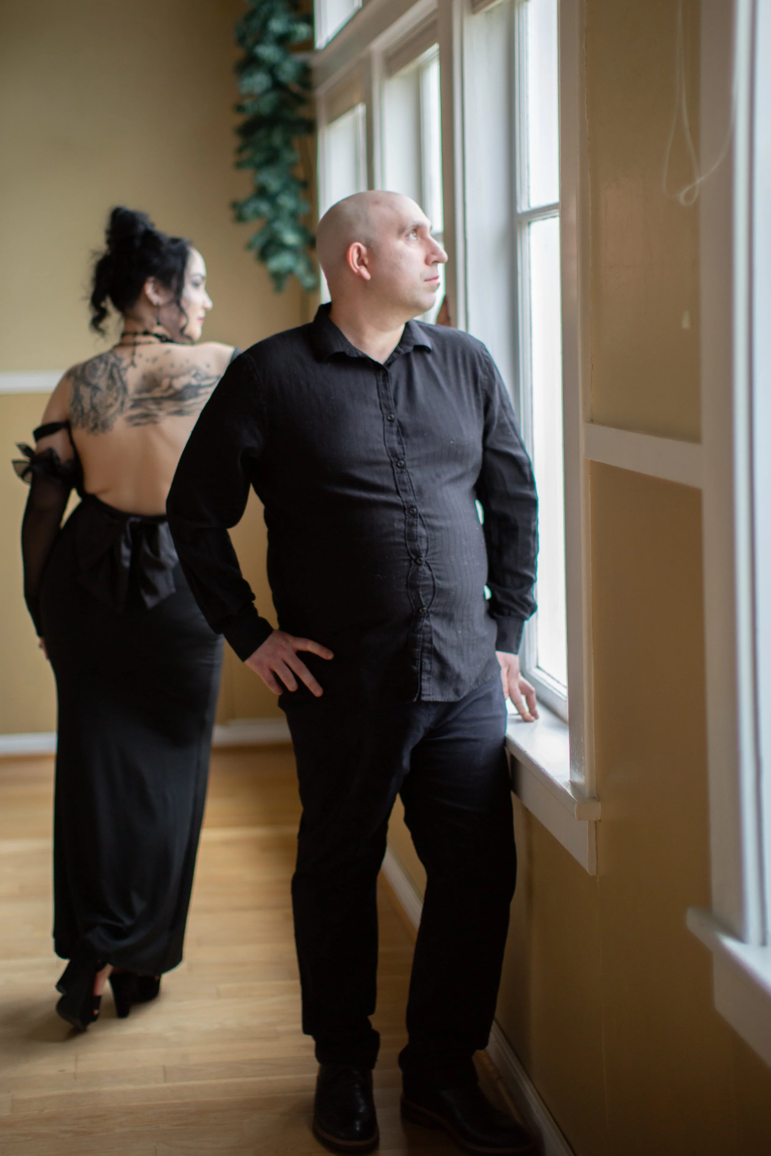 A man with a bald head in a black shirt looking out the window, with a woman in black dress with tattoos on her back and curly black hair standing behind him in a room with cream walls and wooden floors.
