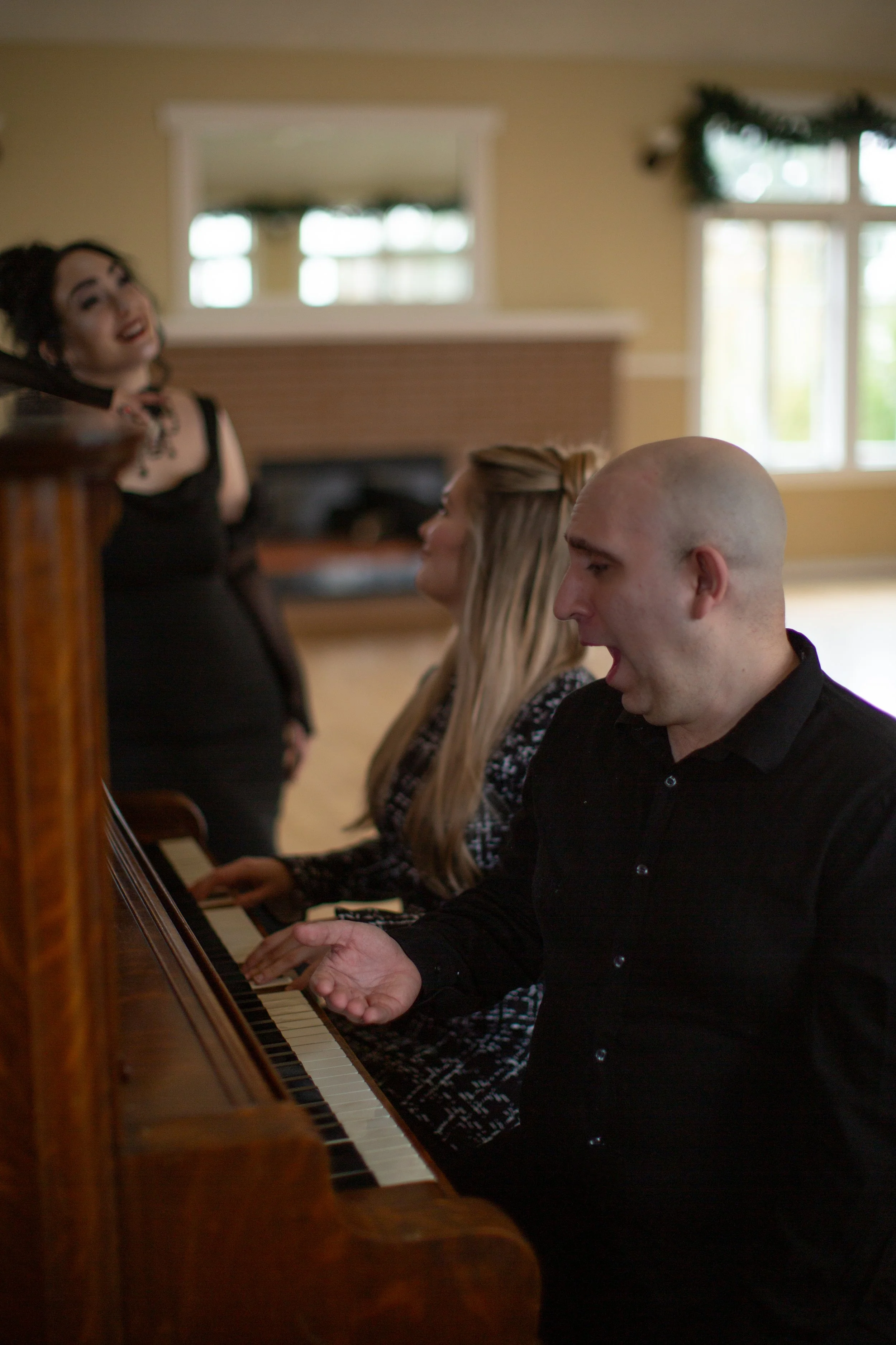 A group of three people, two women and one man, sitting around a piano in a living room, with one woman playing the piano and the others sitting nearby, sharing a moment.