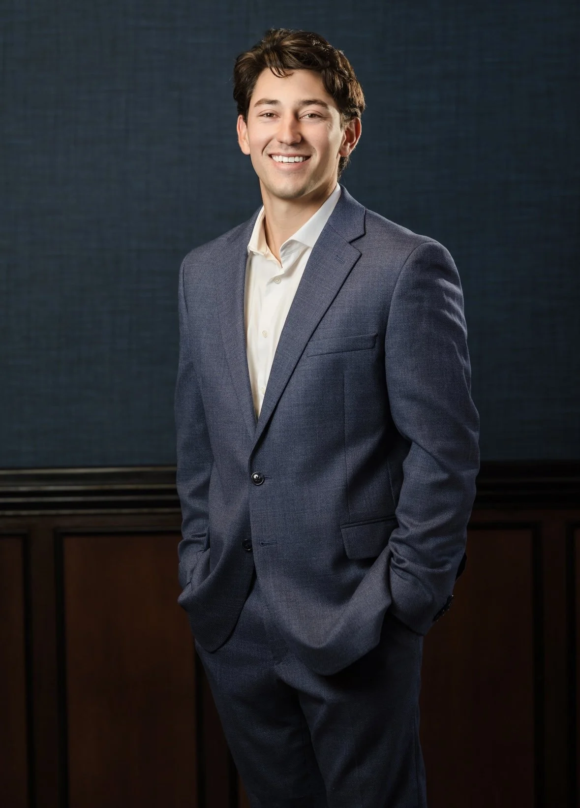 A young man dressed in a gray suit with a white dress shirt, smiling and standing with his hands in his pockets against a dark background.