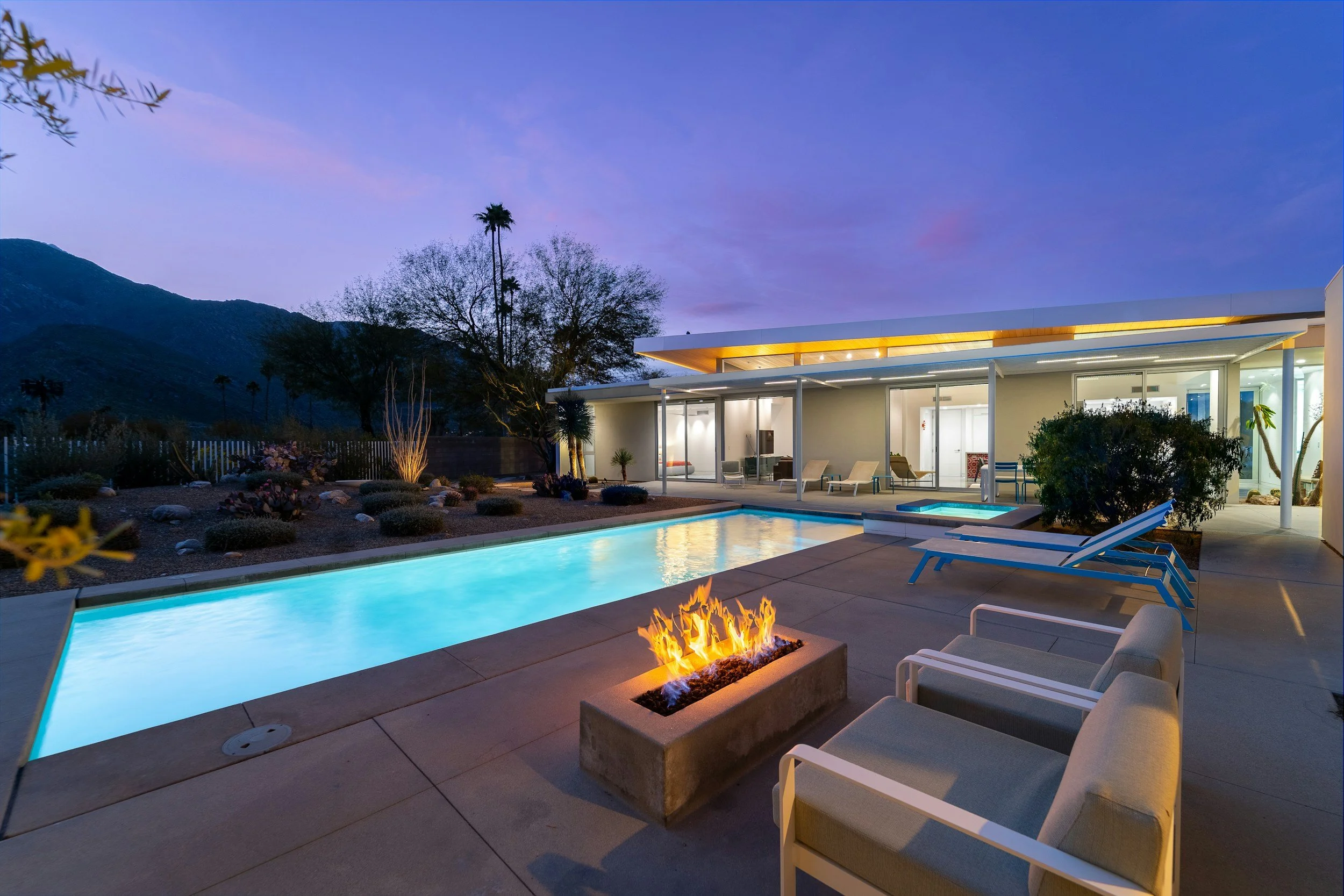 Modern backyard with a swimming pool, outdoor fire pit, lounge chairs, and a glass-walled house at dusk with mountain background.