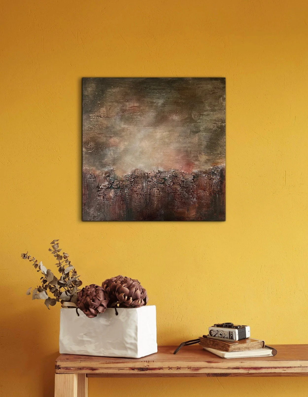 Ein Stillleben mit einer weißen Vase, die Trockenblumen und Eukalyptus enthält, auf einem Holztisch vor einer gelben Wand. Darunter liegt ein Buch und eine kleine Kamera.