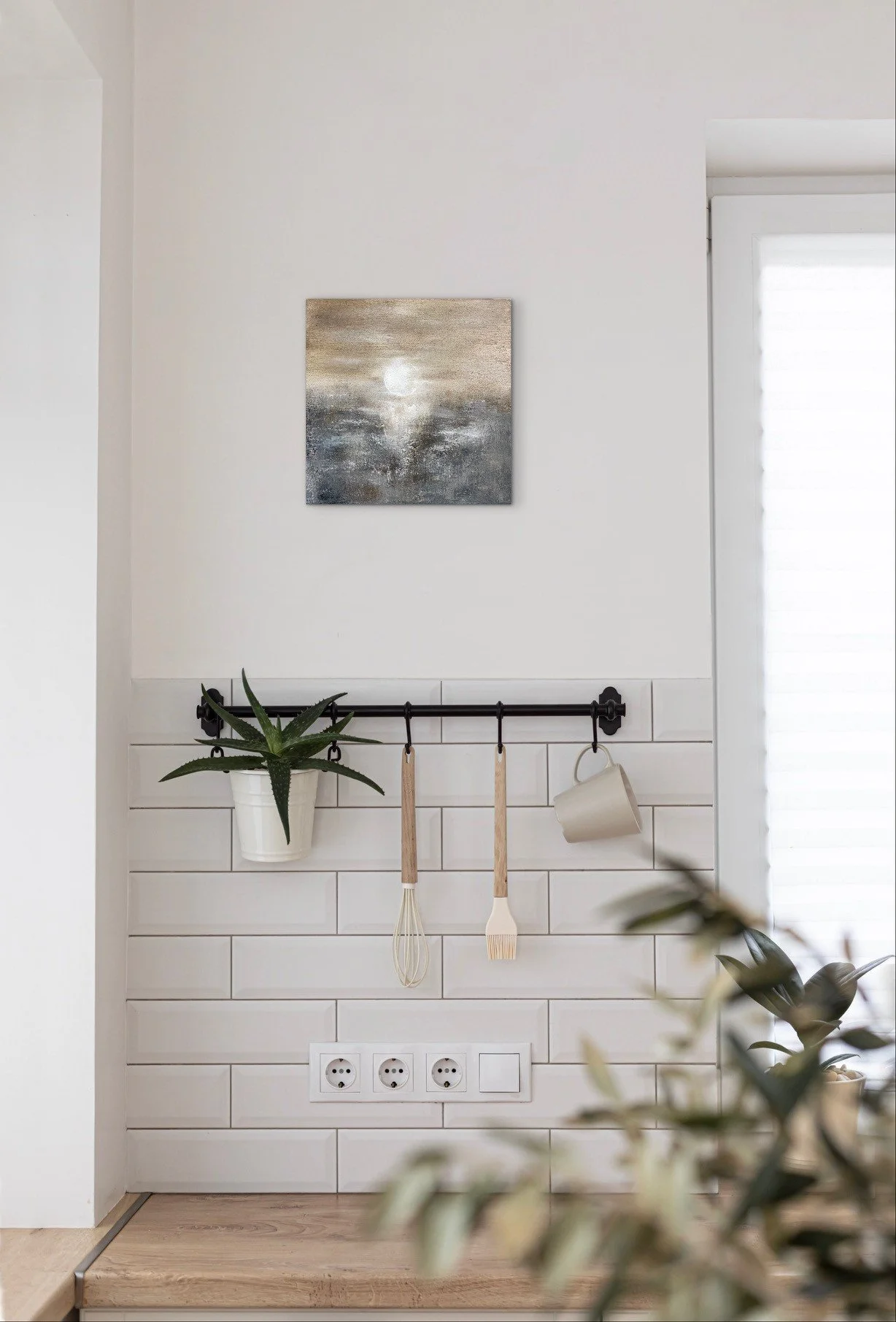 Küchenwand mit weißen Fliesen, einer schwarzen Hakenstange mit Topfpflanze, Küchenutensilien und einer weißen Wandlampe, darüber ein Gemälde mit Mond und Wasser, rechts ein Fenster mit Rollos.