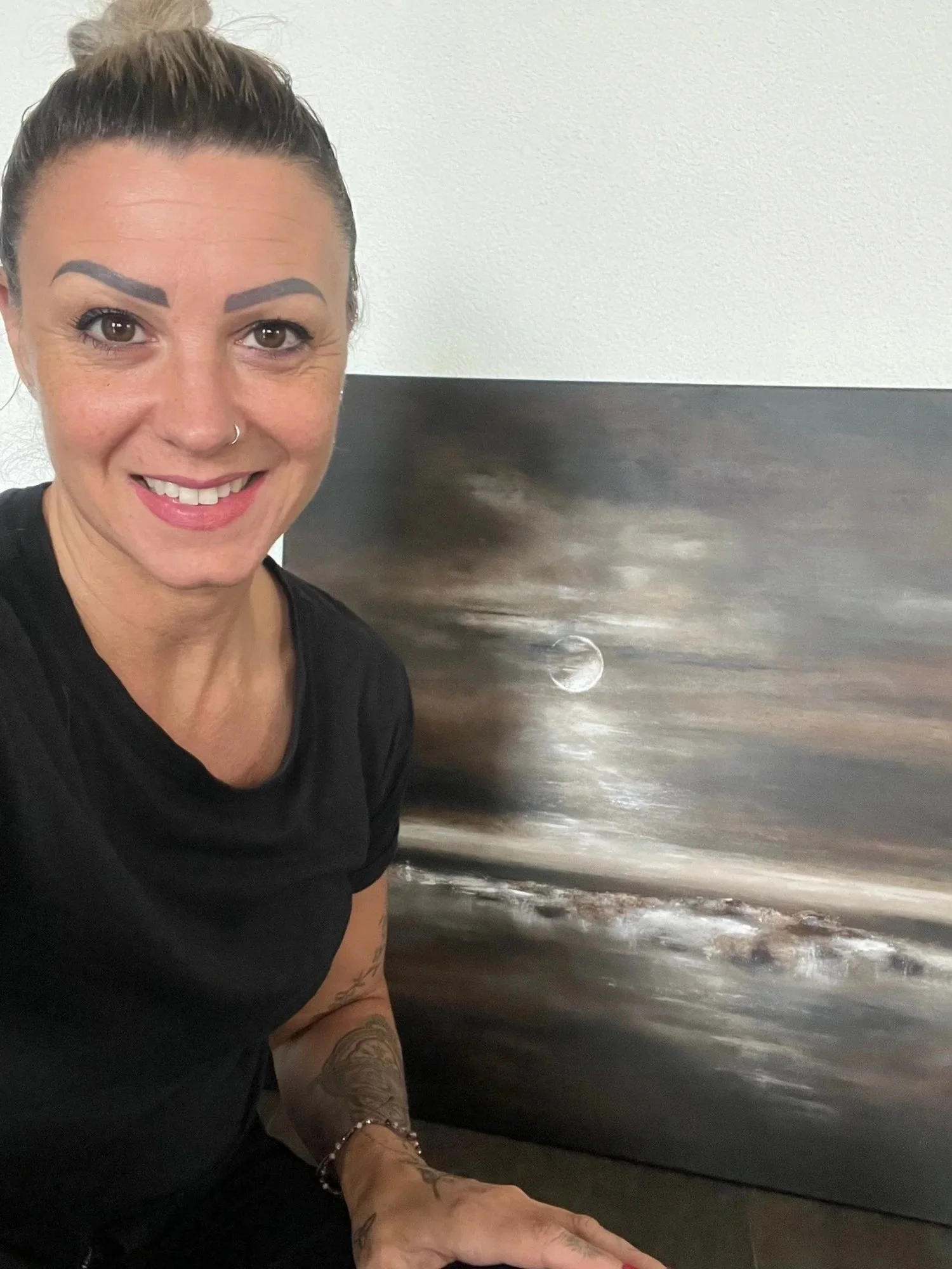 Eine Frau mit dunklem Haarknoten, schwarz Kleidung, tätowierten Armen und Nasenpiercing posiert vor einem Gemälde mit Mond und Himmelsszene.