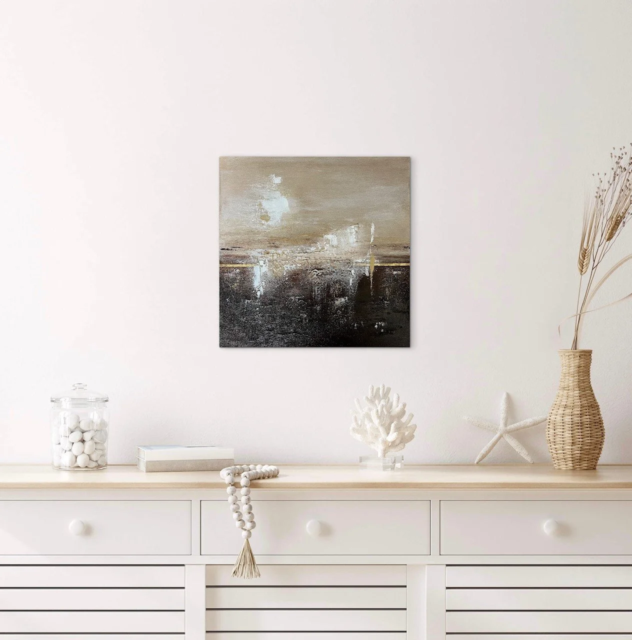 Ein weißer Sideboard mit Dekorationen wie Muscheln, Korallen, eine Glaskare mit weißen Kugeln, ein Naturfarbener Korb mit getrockneten Gräsern und ein kleines Buch. An der Wand darüber hängt ein abstraktes Gemälde in neutralen Tönen.