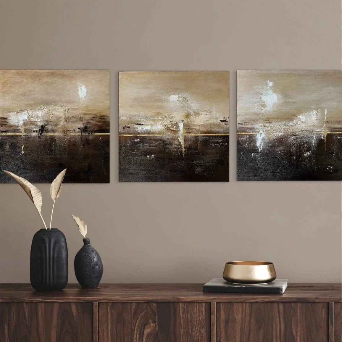Drei abstrakte Gemälde in neutralen Tönen hängen an einer Wand, darunter ein dunkler Holzsideboard mit zwei Vasen, einer großen schwarzen mit Pergamentblättern und einer kleineren schwarzen, sowie einer goldenen Schale auf einem Buch.