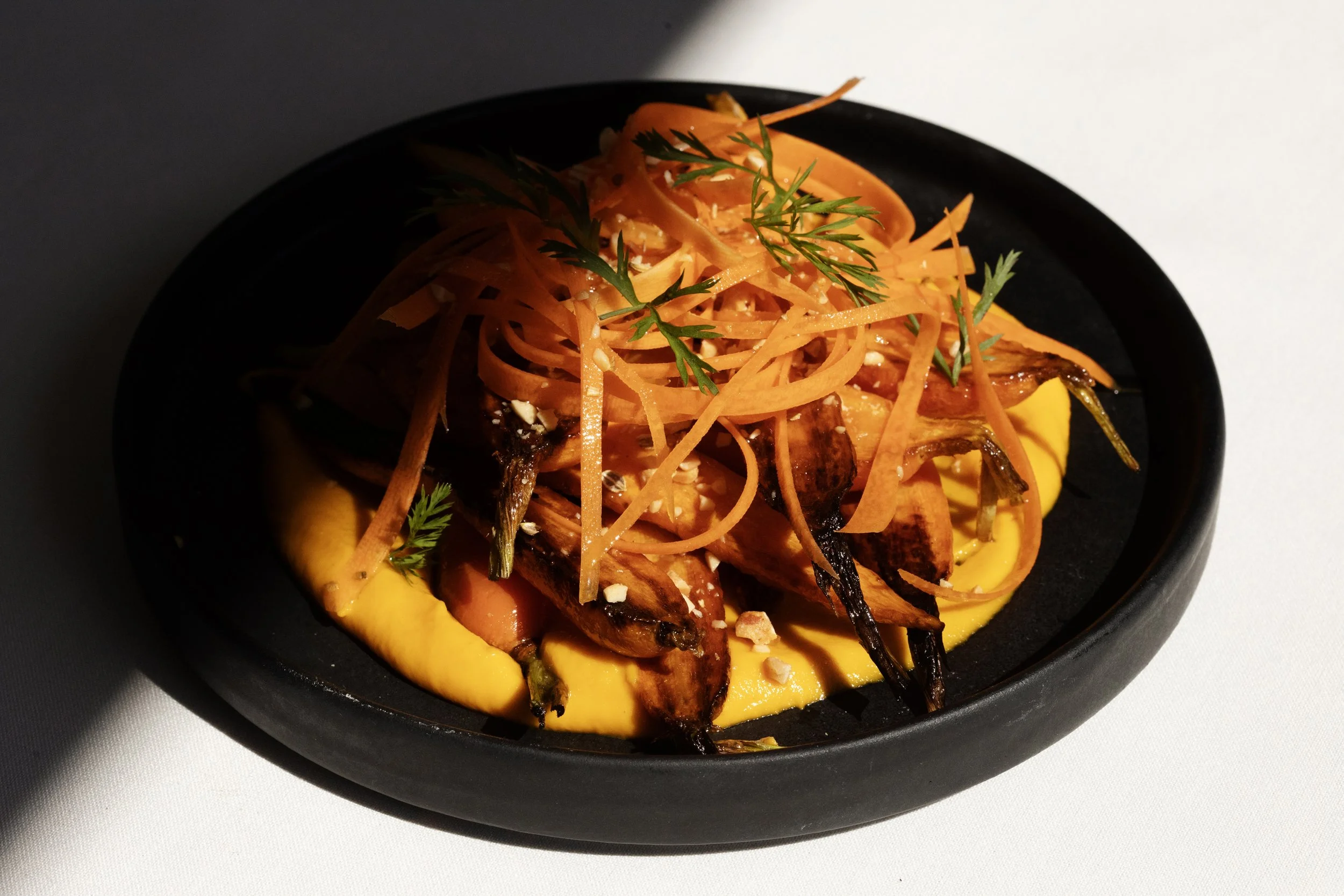 Roasted carrots with carrot ribbons and fresh dill served over a smooth golden purée on a black plate.