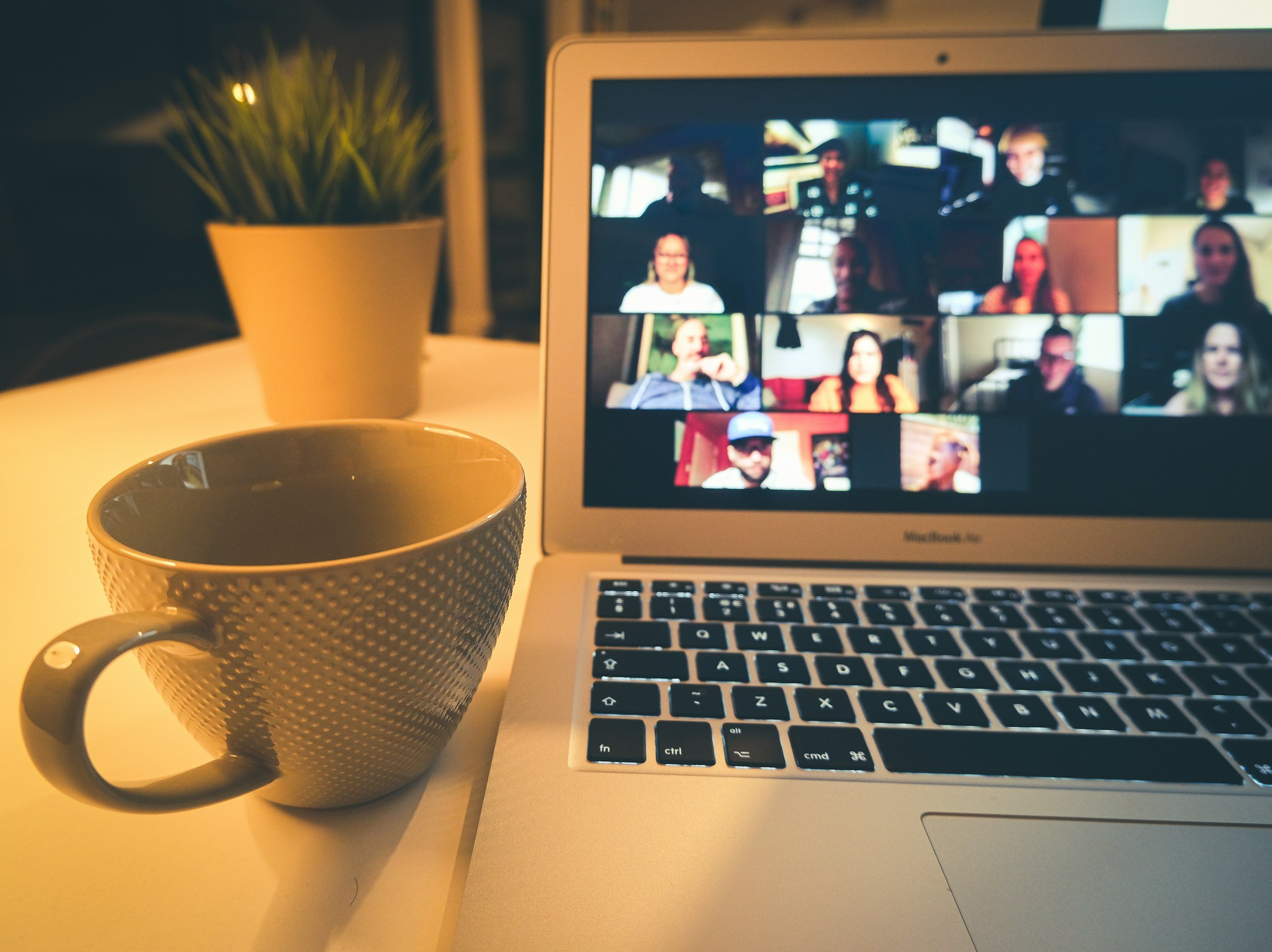 A laptop computer displaying a video call with multiple participants, a white ceramic mug, and a potted plant on a desk with warm lighting.