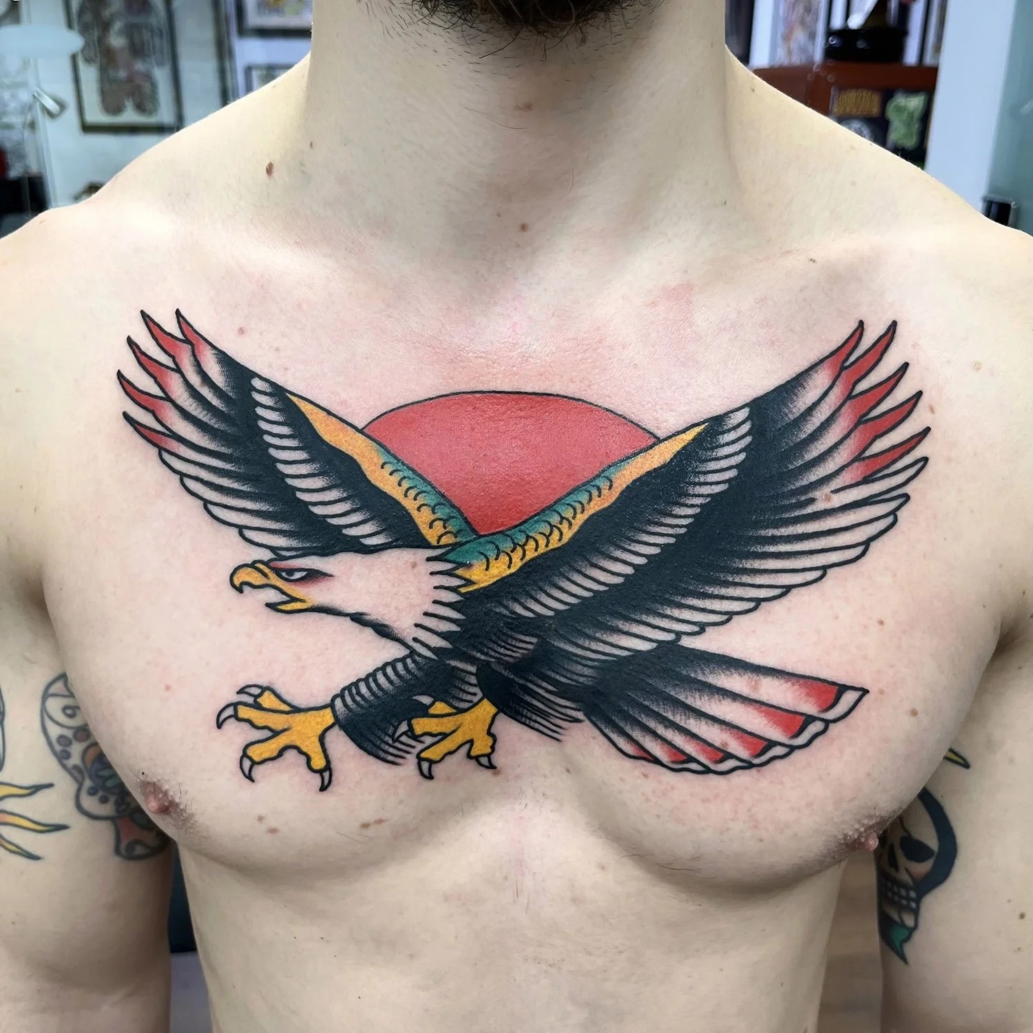 Tattoo of a bald eagle with spread wings, positioned over a red circle on a person's chest, with a tattooed arm visible on the side.
