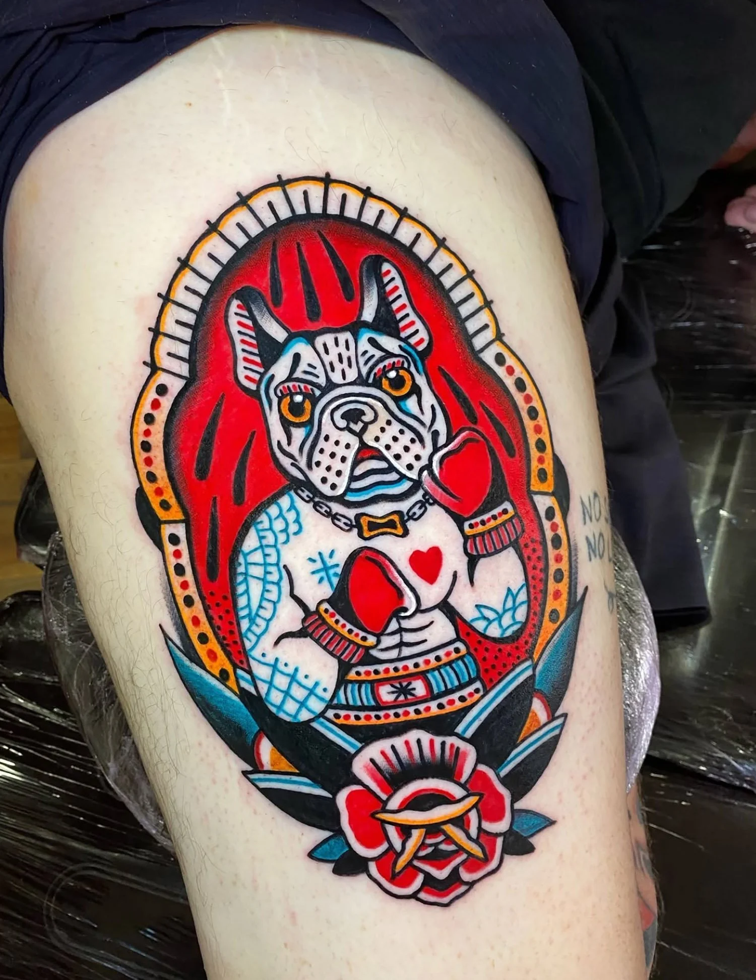 Tattoo of a bulldog with boxing gloves in a traditional style, surrounded by a decorative frame and a red rose at the bottom.