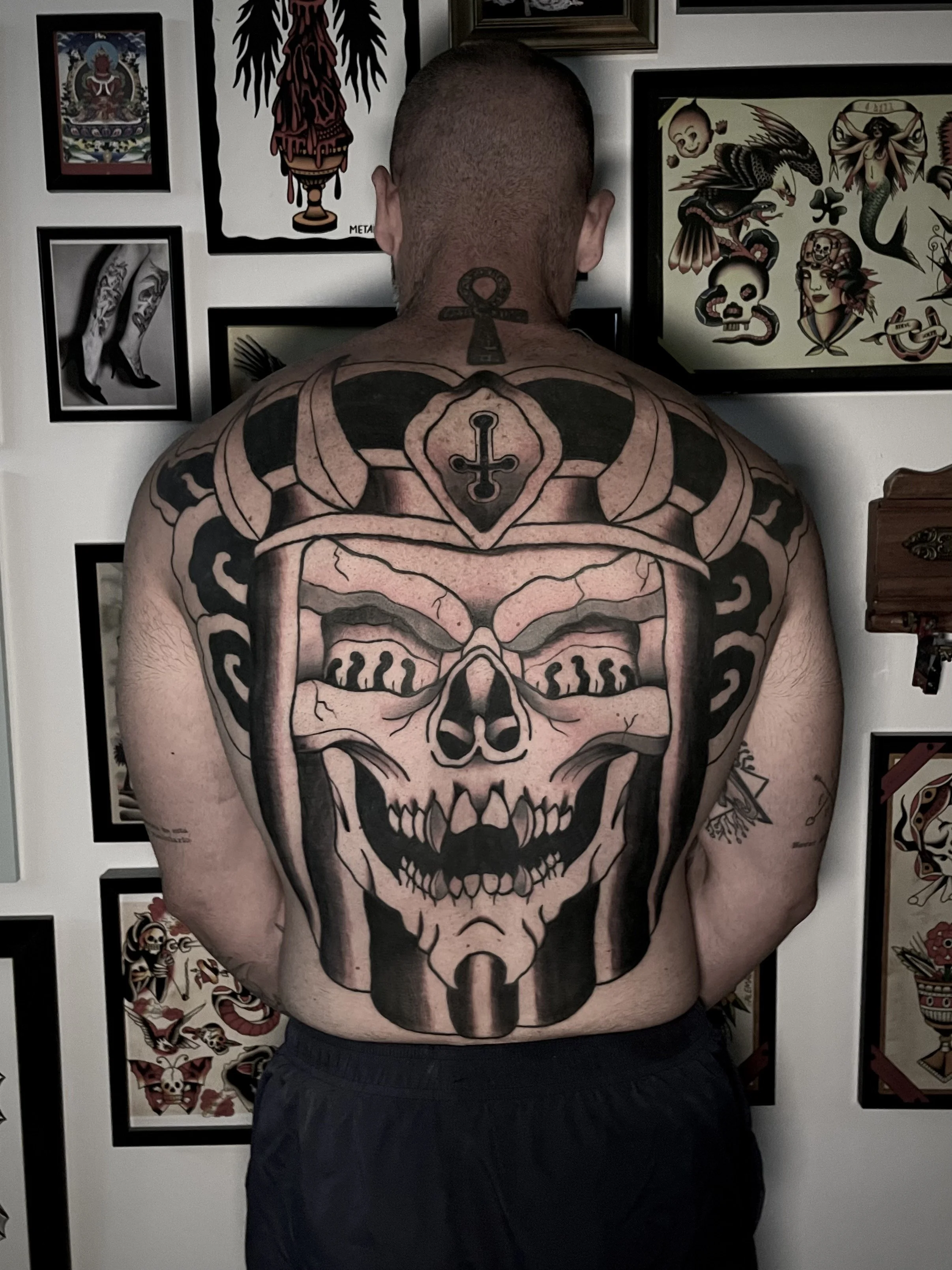 Tattooed man with large skull and helmet tattoo on his back, standing in front of a wall decorated with tattoo and art-related framed pictures.