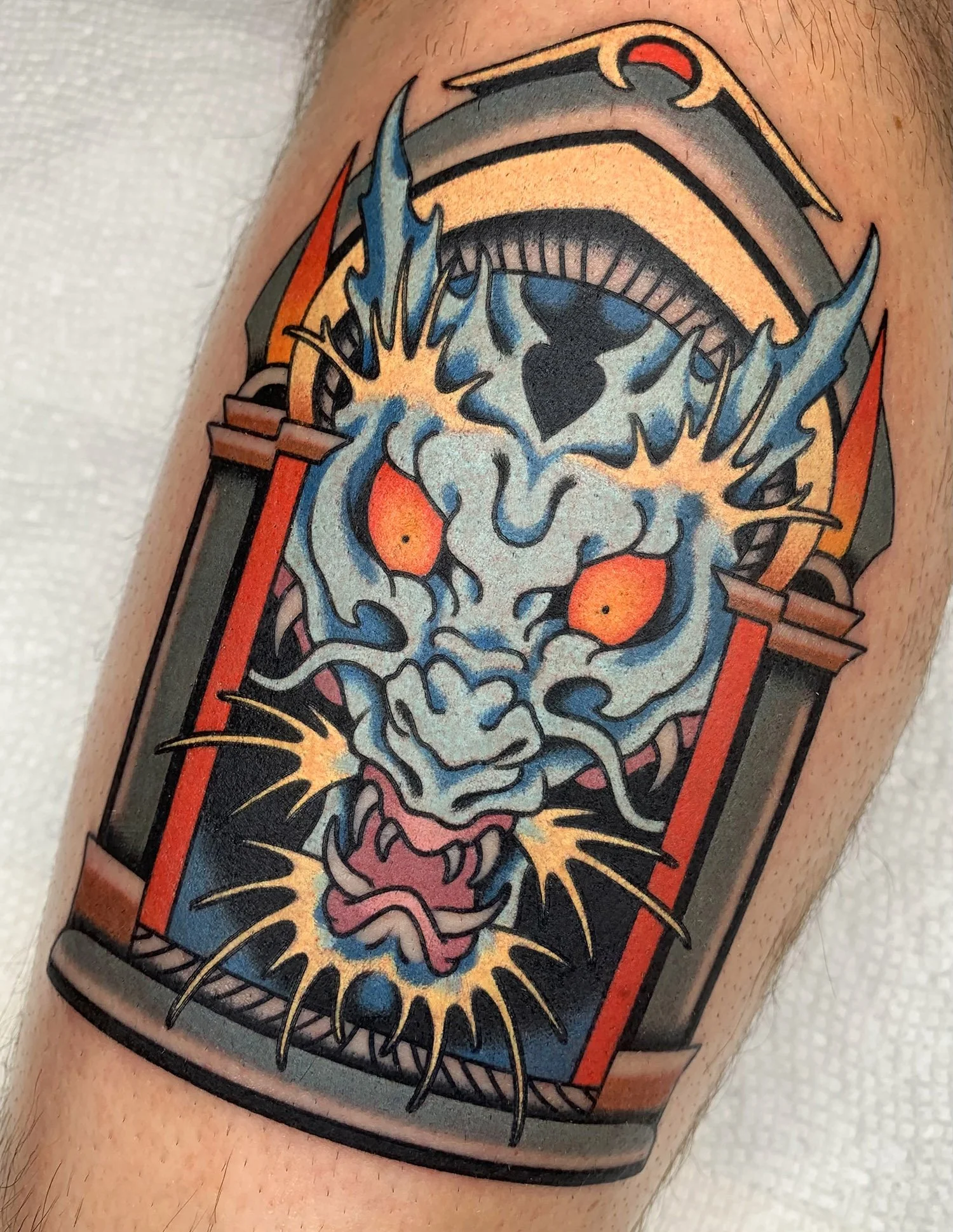 Tattoo of a fierce dragon skull with fiery eyes inside a decorative arch with bold black outlines, featuring vibrant colors like blue, orange, and yellow.