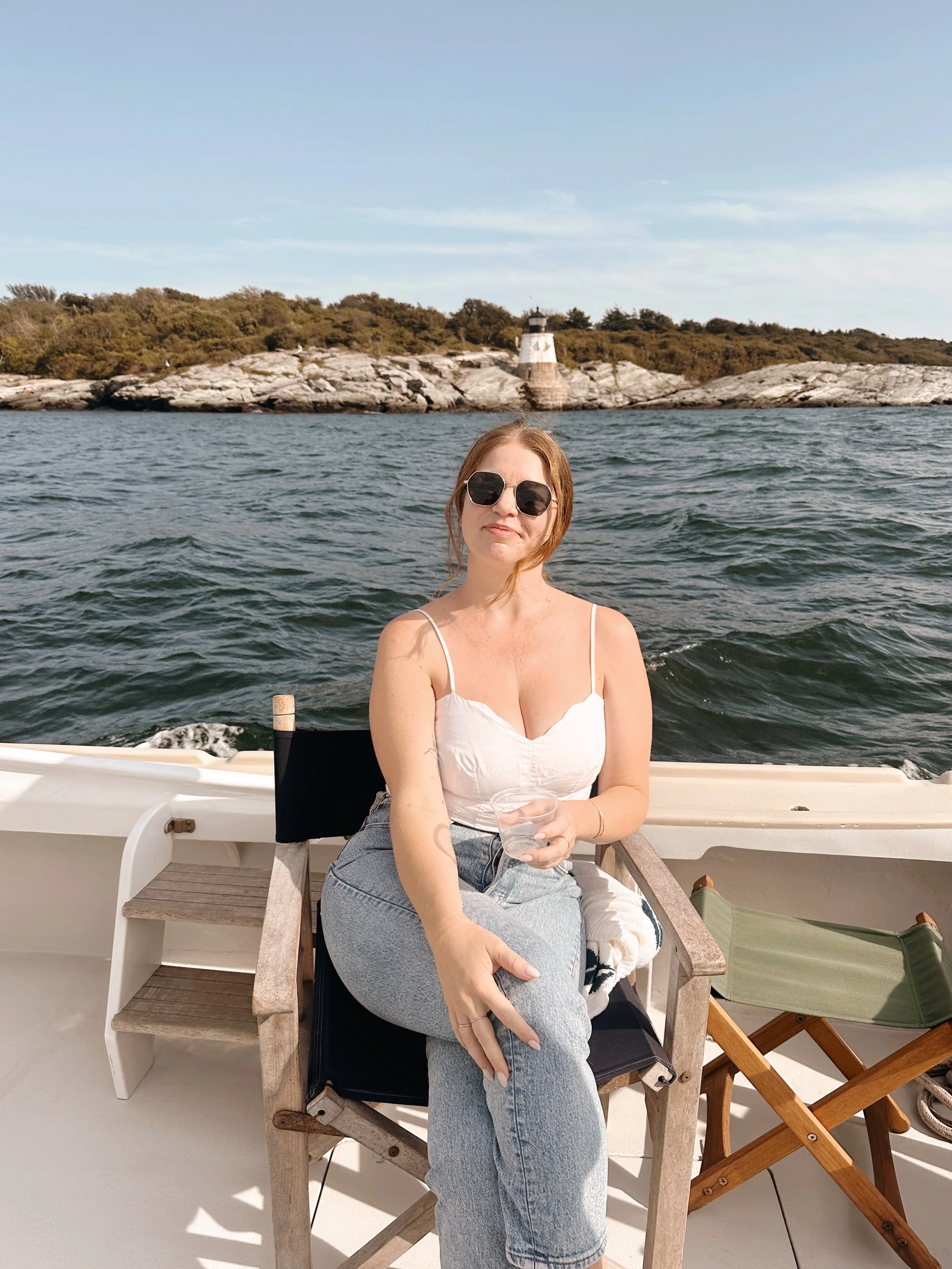 A woman sitting on a boat, wearing sunglasses and a white tank top, holding a glass of drink, with water, rocky shoreline, trees, and a lighthouse in the background.