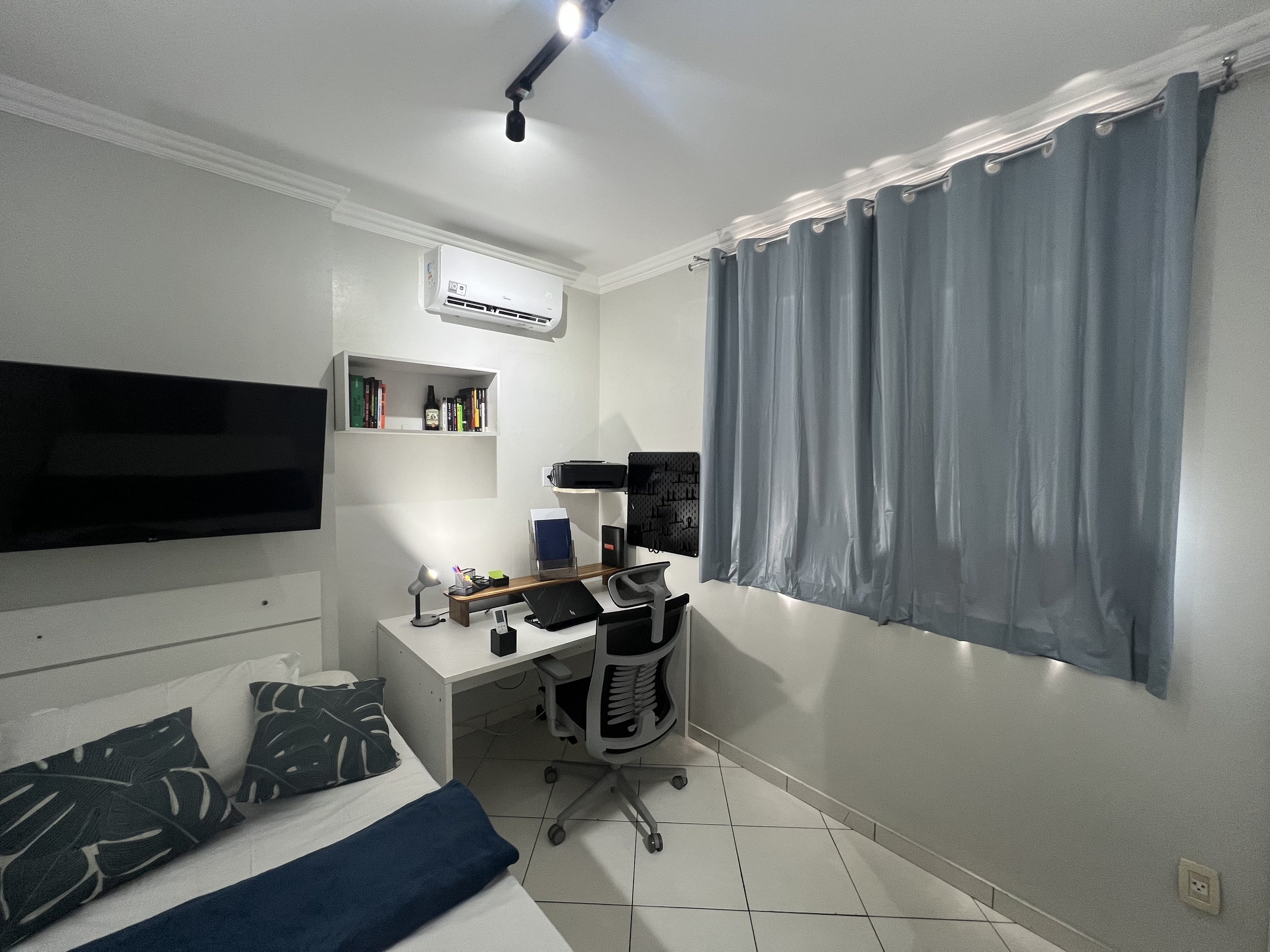 A small bedroom with a bed, a desk, a chair, a television mounted on the wall, a bookshelf, a window with blue curtains, and an air conditioner above the bookshelf.