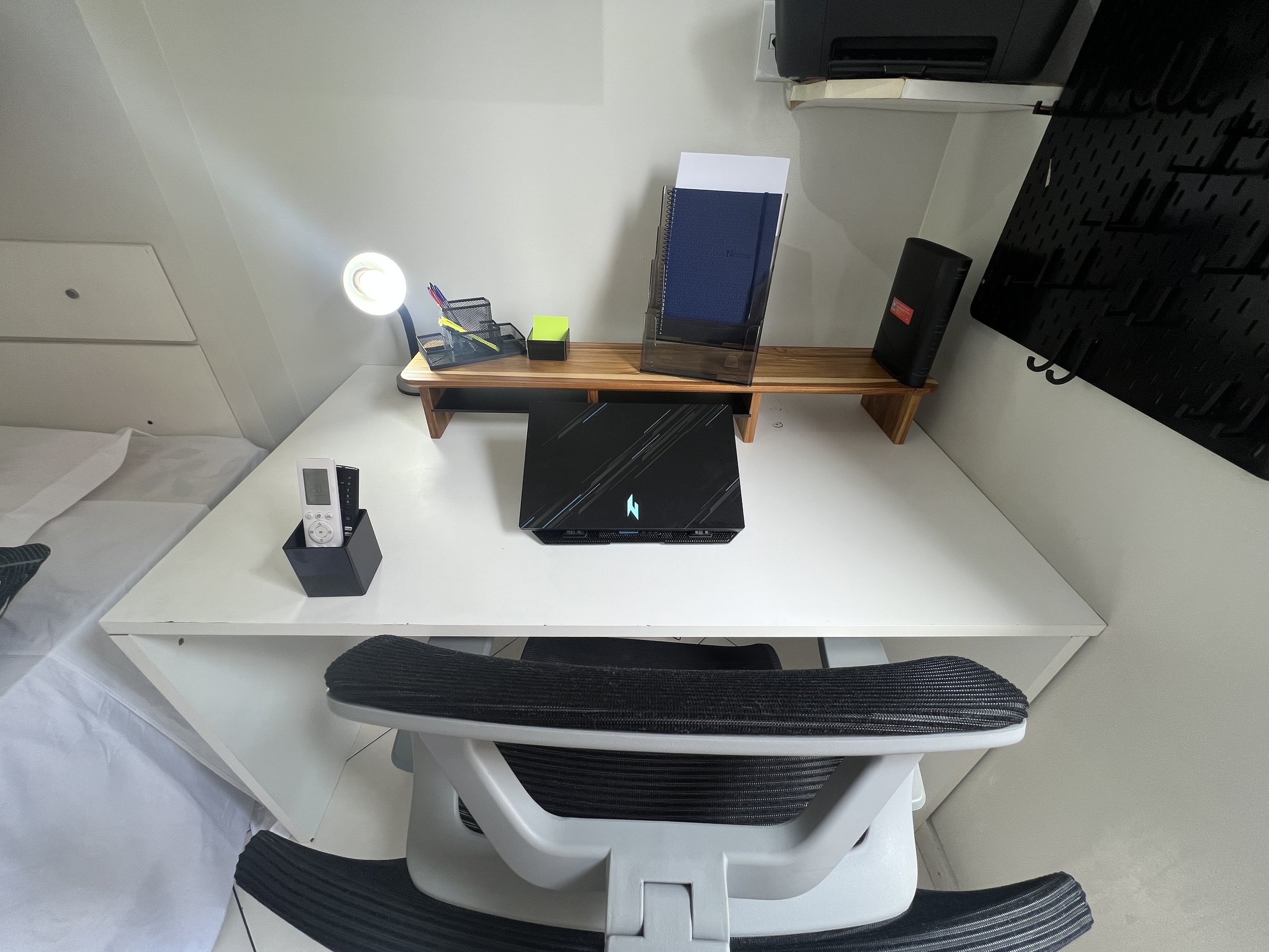 Office desk with a white surface and black office chair. On the desk, there is a black laptop, a small black pen holder, a white landline phone, and a wooden shelf behind the desk with a blue notebook, a neon green item, and electronic devices. A wall-mounted TV is visible above the desk.