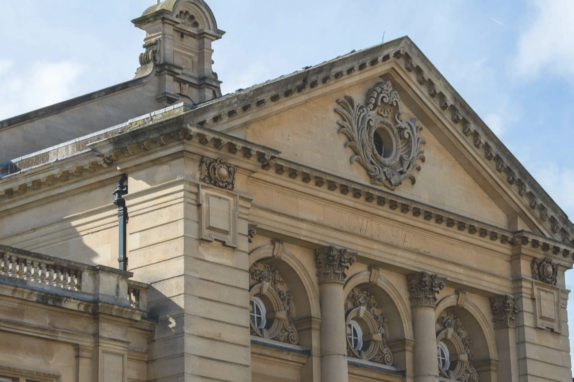 Close-up of a classical style building's upper facade with ornate decorations, columns, and a clockless round window.