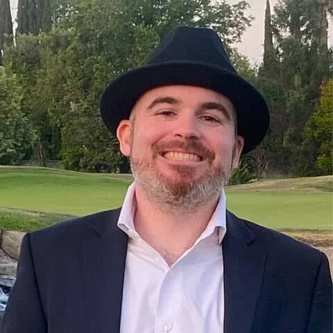 A man smiling outdoors, wearing a black fedora hat, white dress shirt, and dark blazer, with trees and grass in the background.