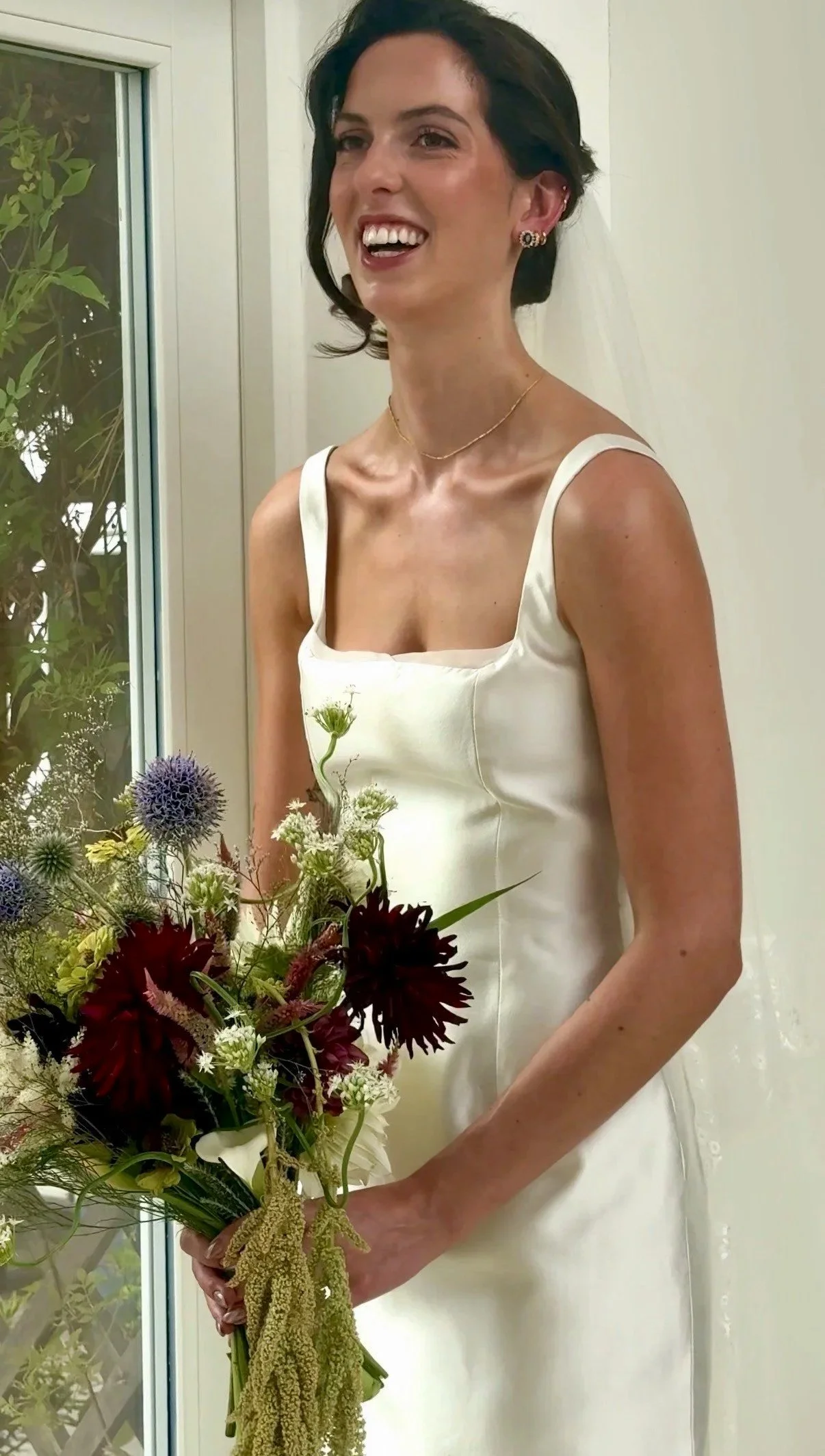 A bride laughs while she stands near the window. Her makeup looks radiant and she wears a berry coloured lip. Her hair is in a low bun with soft curls framing her face.