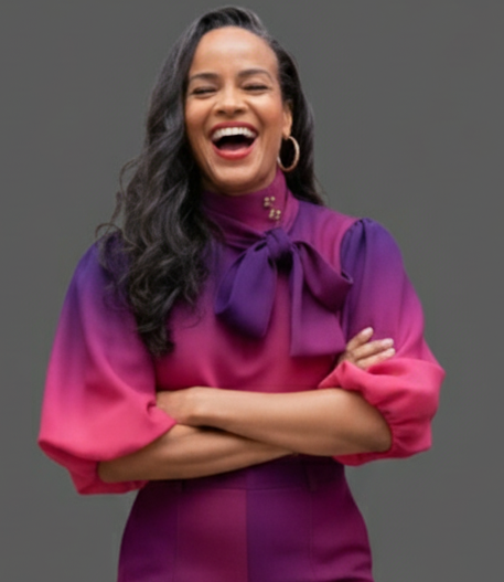 A woman with dark hair, smiling and laughing with arms crossed, wearing a purple and pink dress with a bow at the neck and hoop earrings.