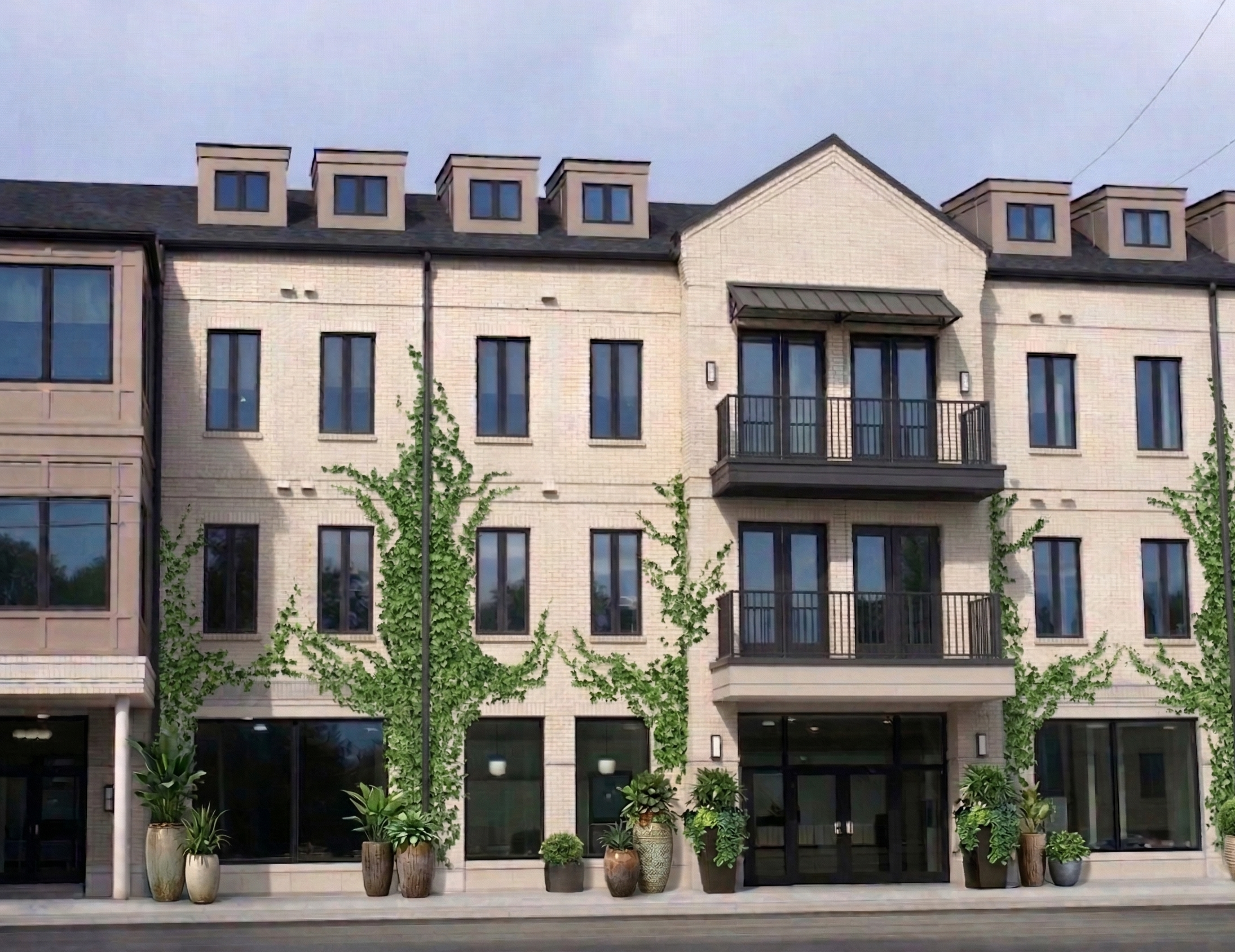 Modern multi-story residential building with brick facade, large windows, and balconies, surrounded by potted plants and small trees, under a cloudy sky.