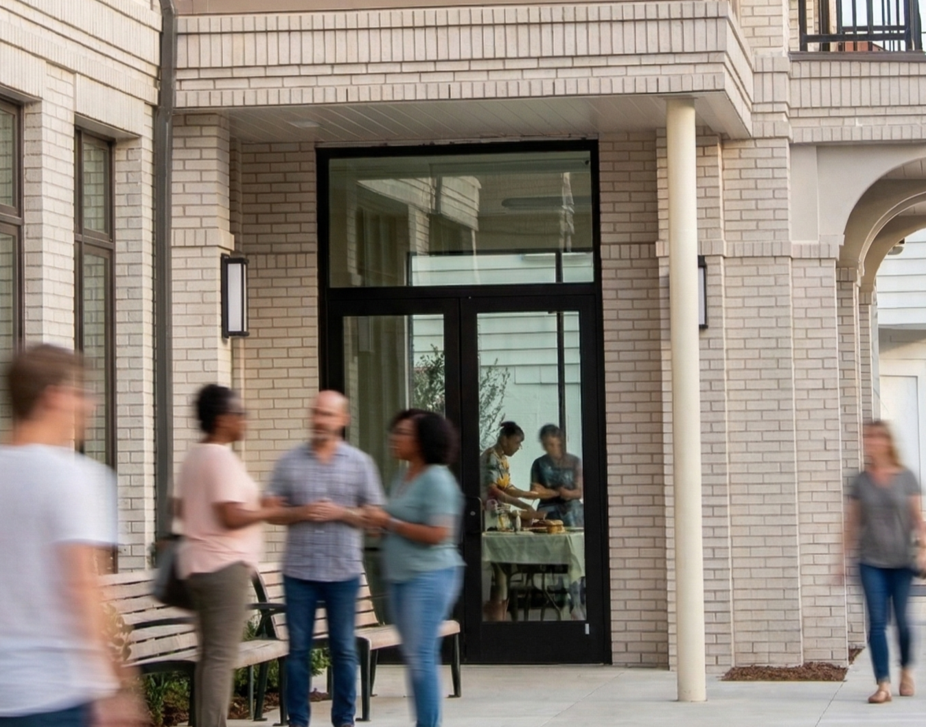 People socializing outside a building with large windows and brick walls, with some inside visible through the glass.