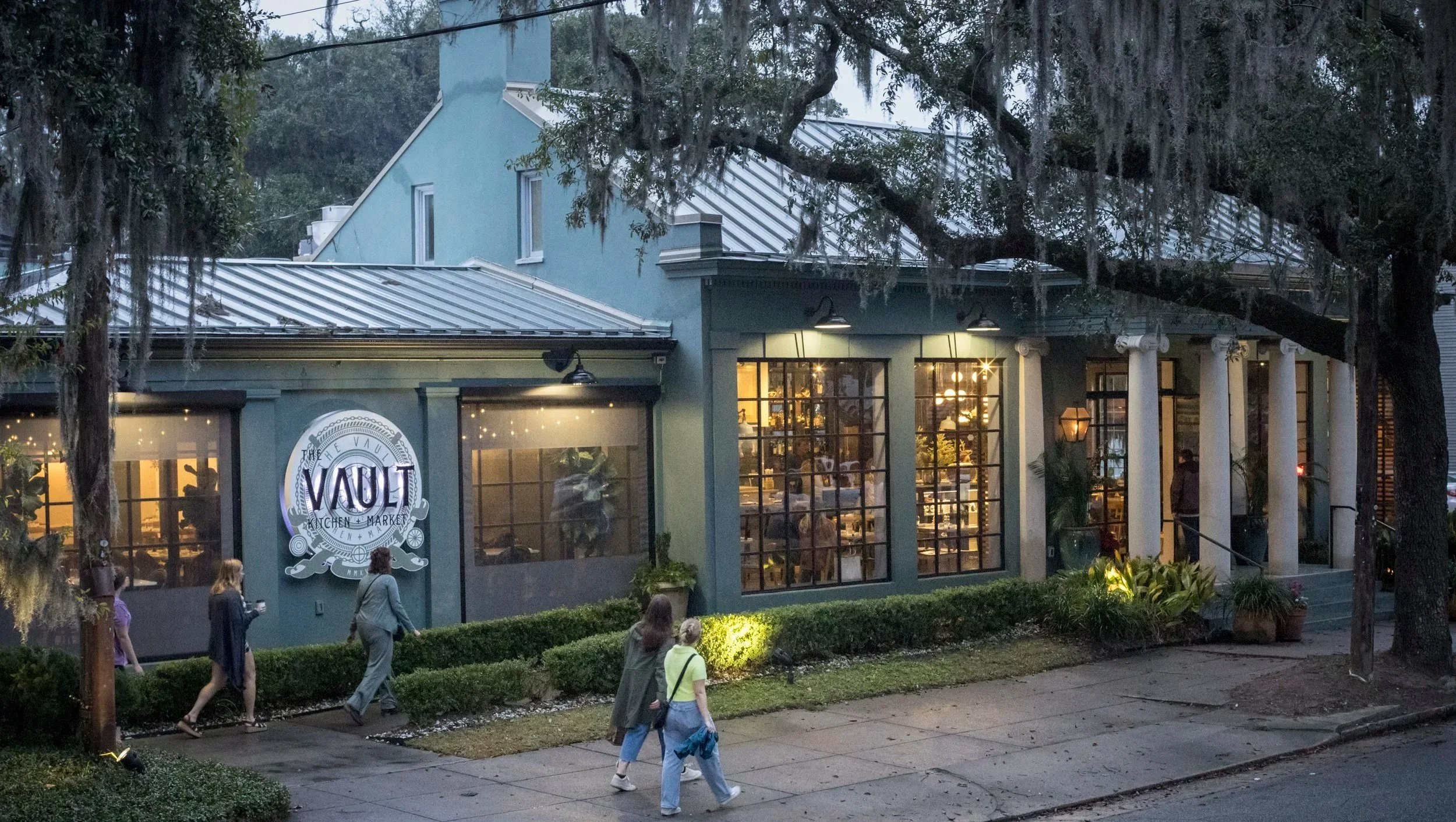 A restaurant called 'The Vault' with large glass windows, white columns at the entrance, and a sign on the front door. People are walking on the sidewalk outside, and warm indoor lighting is visible through the windows. Large trees are in the foreground.