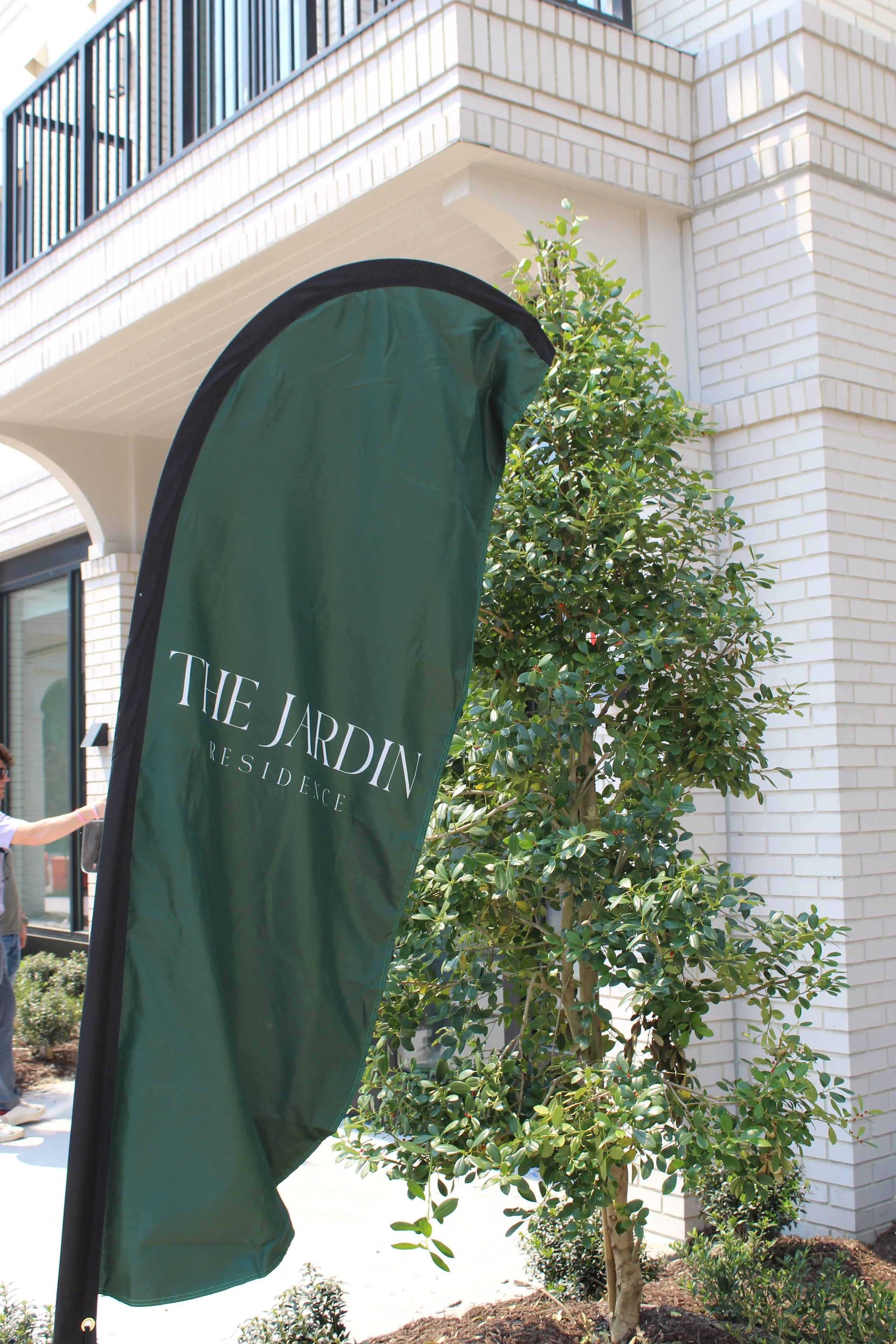 Green promotional banner for 'The Jardin Residence' outside a modern residential building with white brick exterior, trees, and plants.