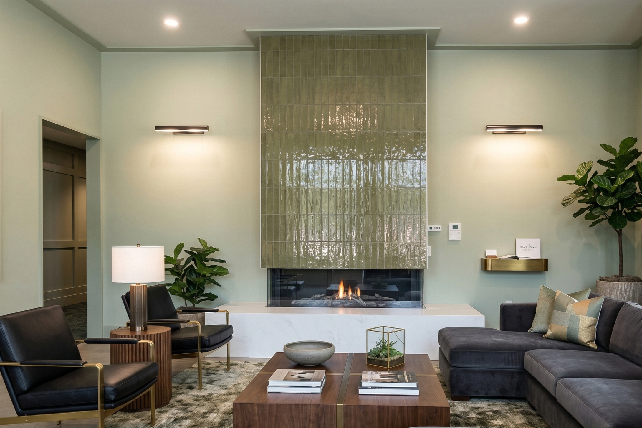 Modern living room with a fireplace, green tile wall, black leather chairs, a dark gray sofa with multicolored pillows, a wooden coffee table with books and a glass terrarium, side table with lamp, green plants, and soft lighting.