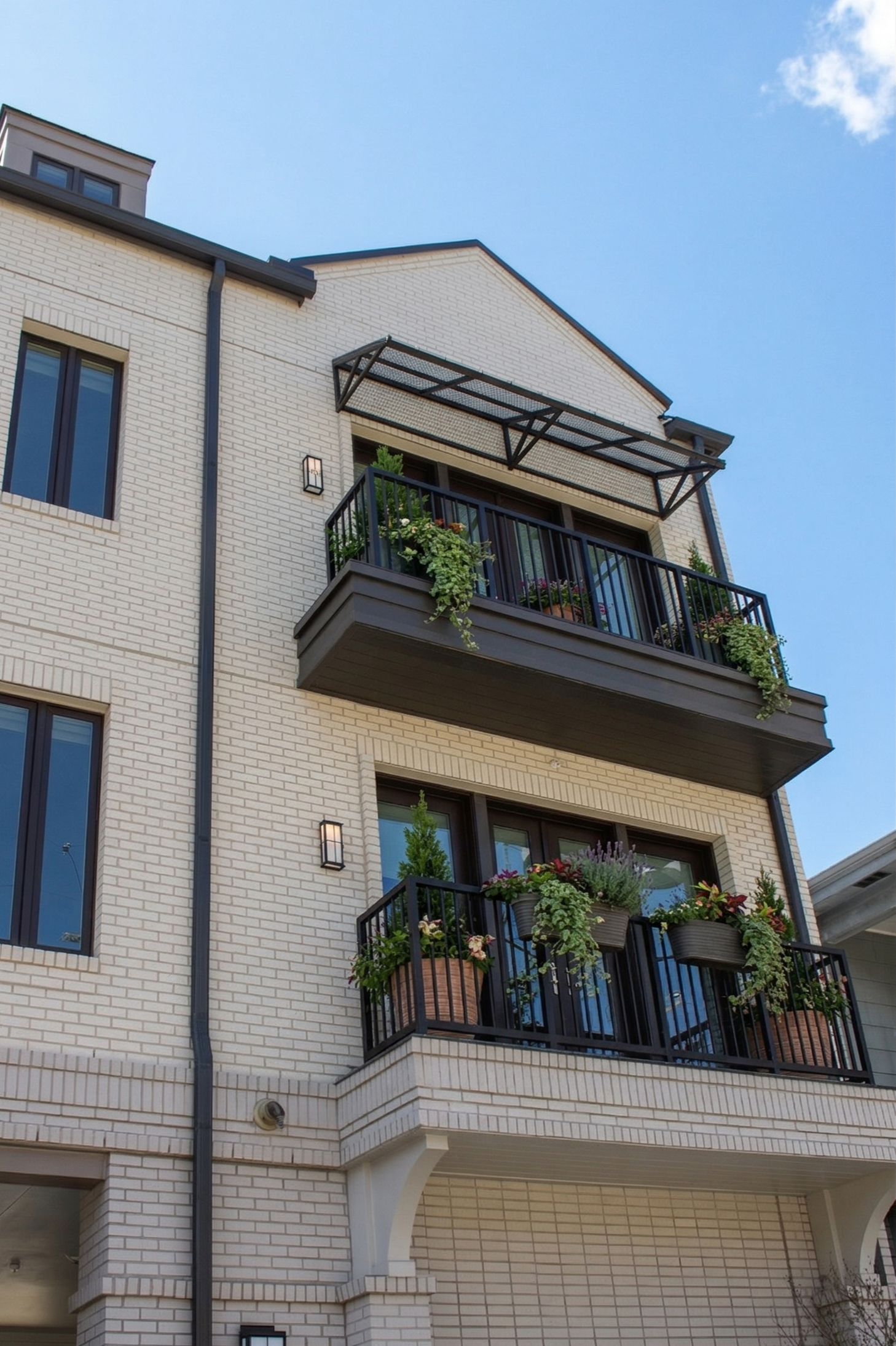 Front view of a three-story apartment building with brick exterior, black balconies with potted plants, and a clear blue sky.