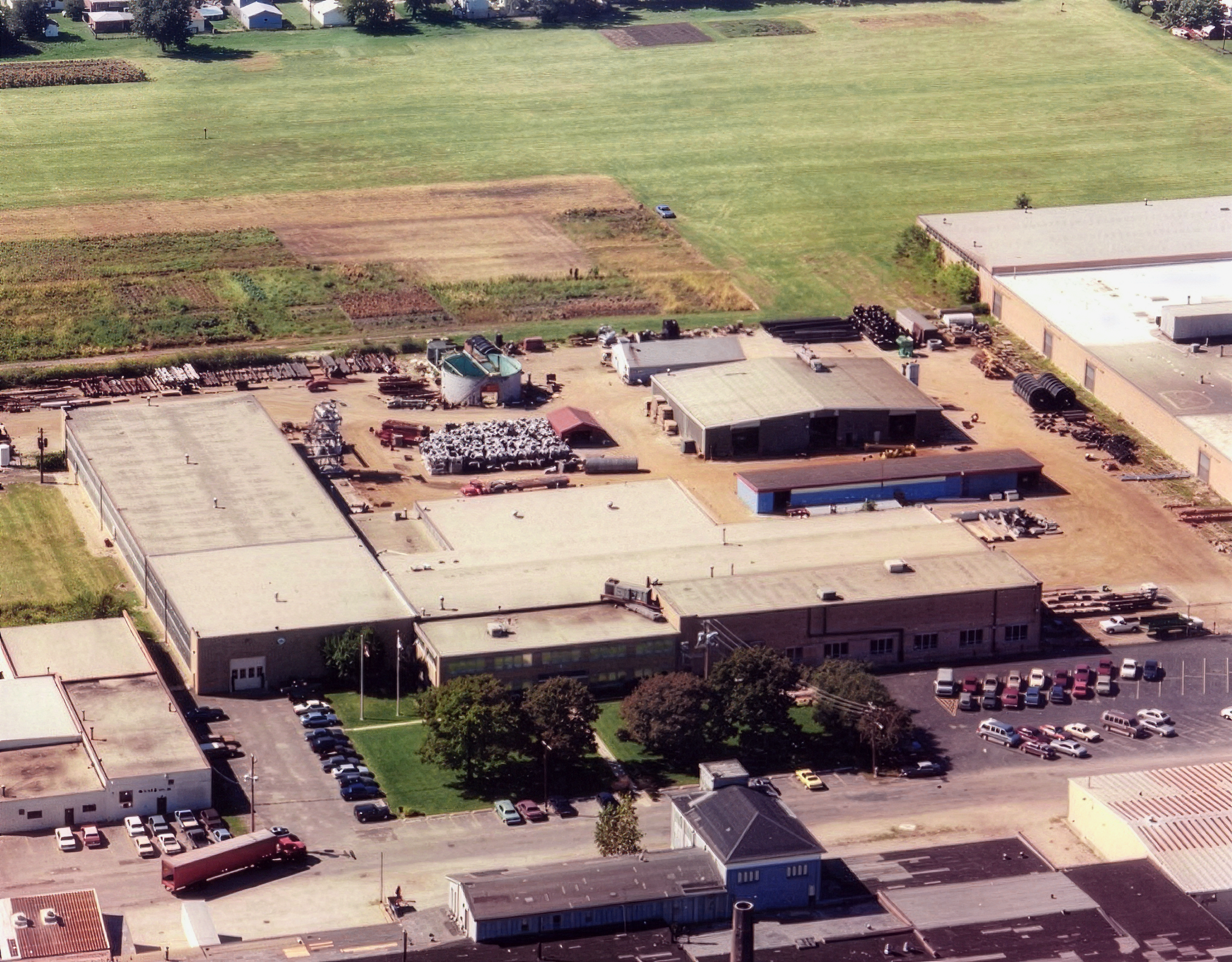 An aerial view of Walker Process Equipment manufacturing facilities with several large flat-roofed buildings, parking lots filled with cars, and nearby farmland with green fields and some cultivated plots.