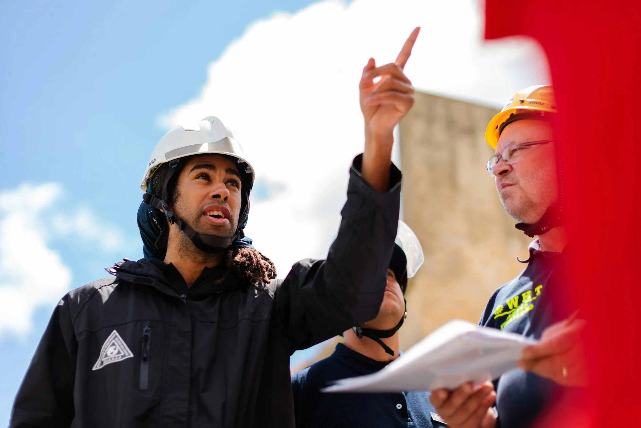 Job site workers wearing safety helmets, one man with a white helmet pointing and talking to a man with a yellow helmet holding papers, outdoors on a sunny day.