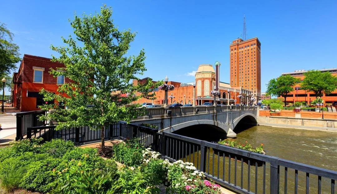Aurora Illinois cityscape with a stone bridge over a river, surrounded by colorful buildings, trees, and flowers under a bright blue sky.