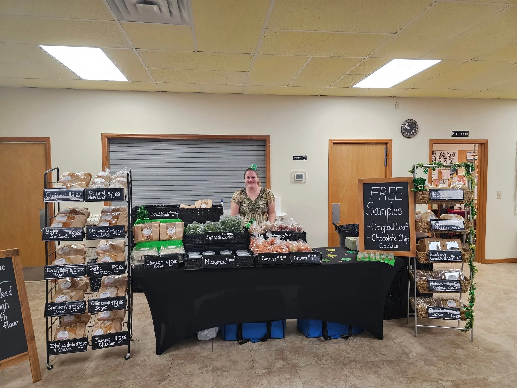 S.J. Organic Farms team member selling fresh sourdough, bagels, muffins, and produce at an indoor farmers market stand