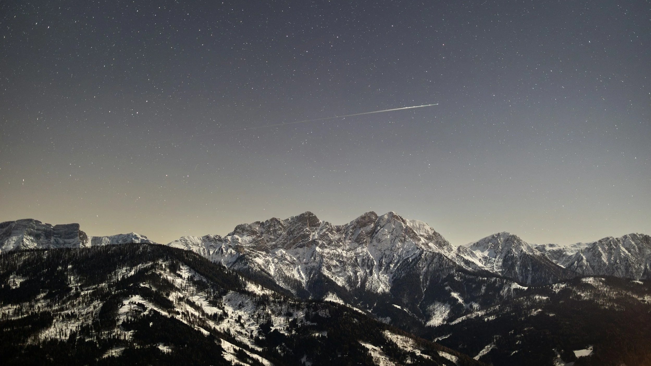 Nighttime view of snow-covered mountain range under a starry sky with a visible airplane trail.