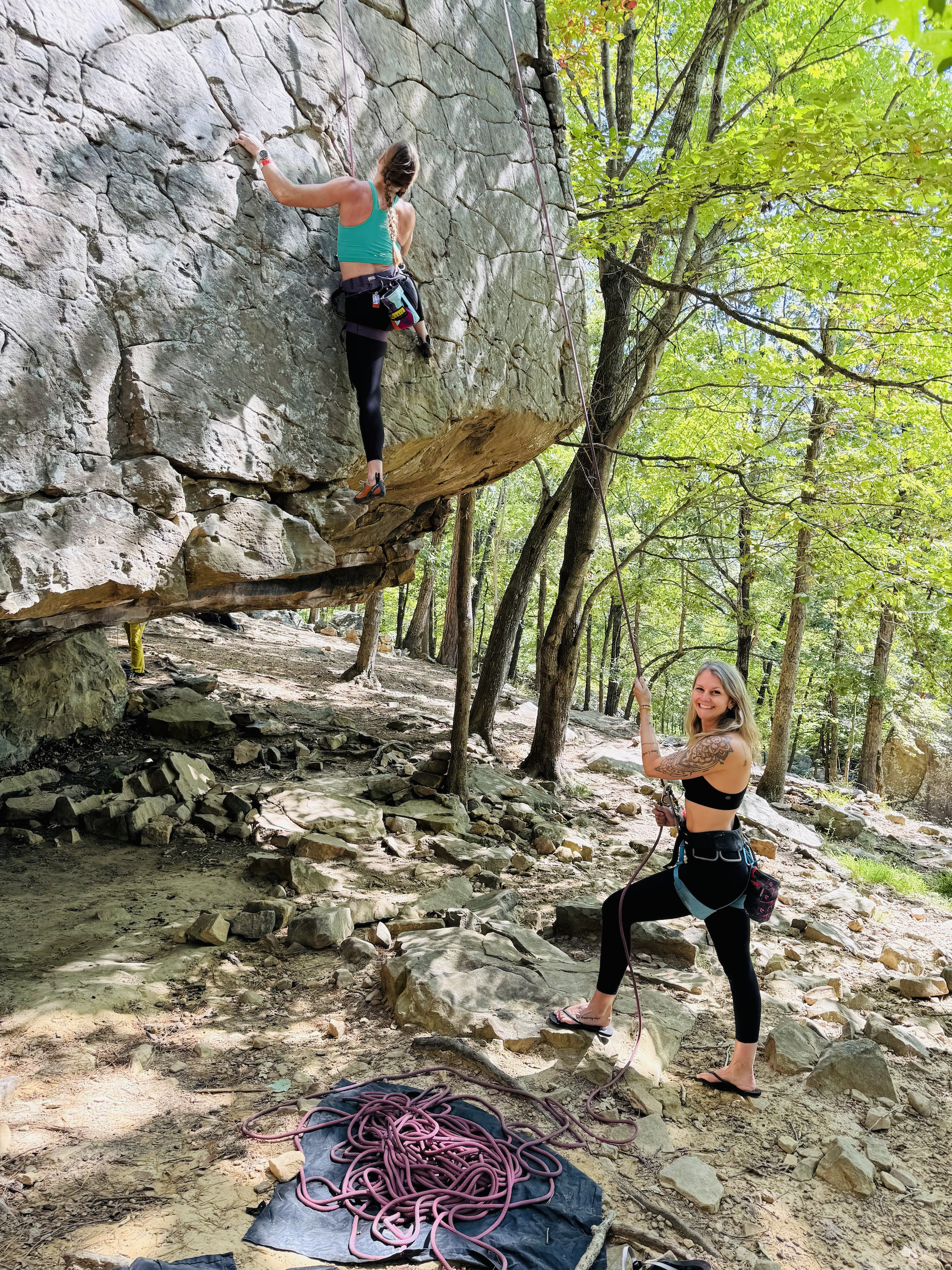 Two women rock climbing outdoors; one woman is climbing a large rock face wearing a turquoise sports bra and black leggings, while the other woman, standing on the ground holding a rope, is wearing a black sports bra and black leggings, smiling at the camera. There is a tangled climbing rope on the ground and trees in the background.